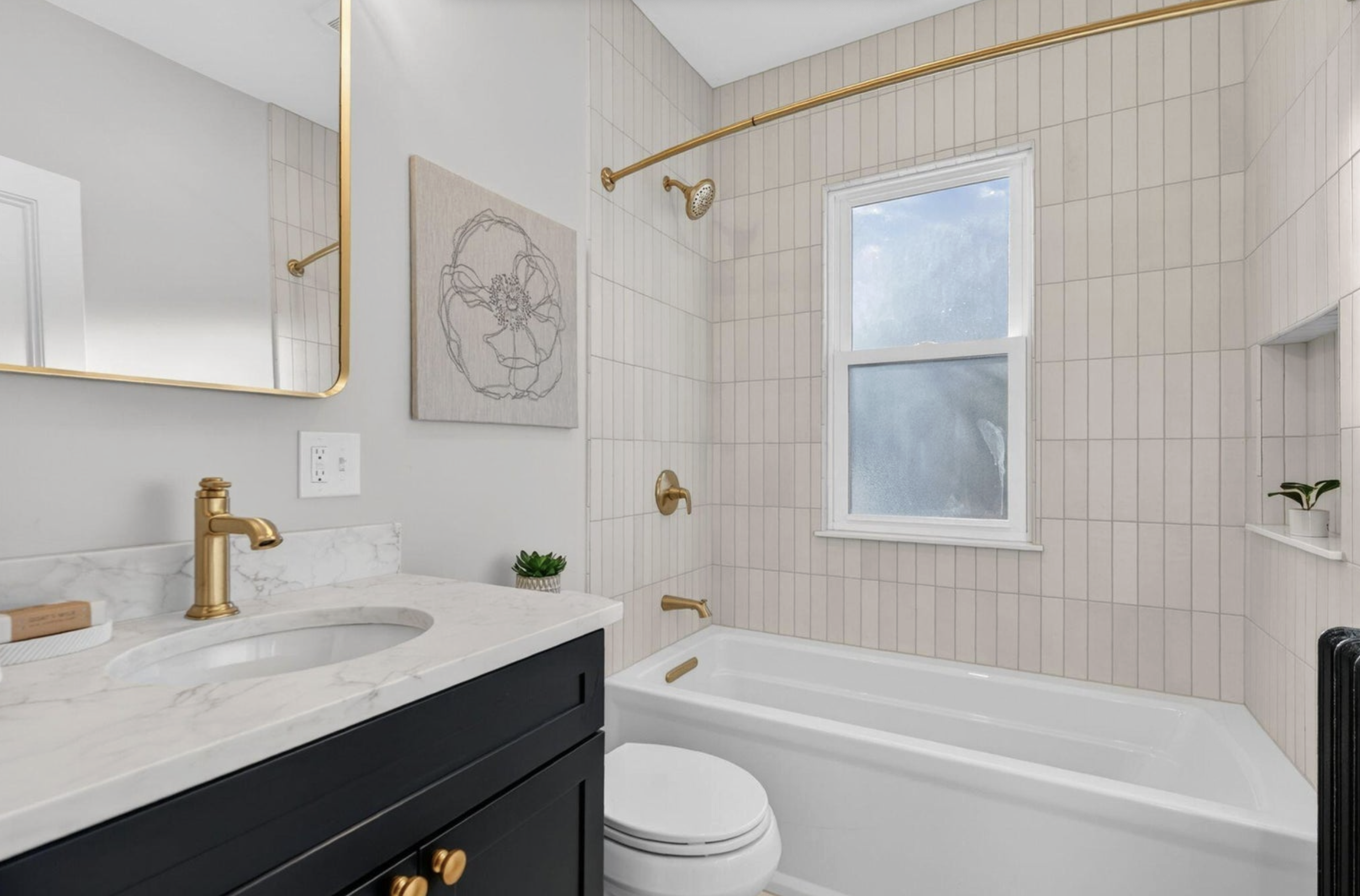 A modern bathroom featuring a white bathtub, a black vanity with gold fixtures, a mirror with a gold frame, artwork on the wall, and a window with frosted glass. There are gold accents on the shower curtain rod, faucet, and curtain hooks, and a small