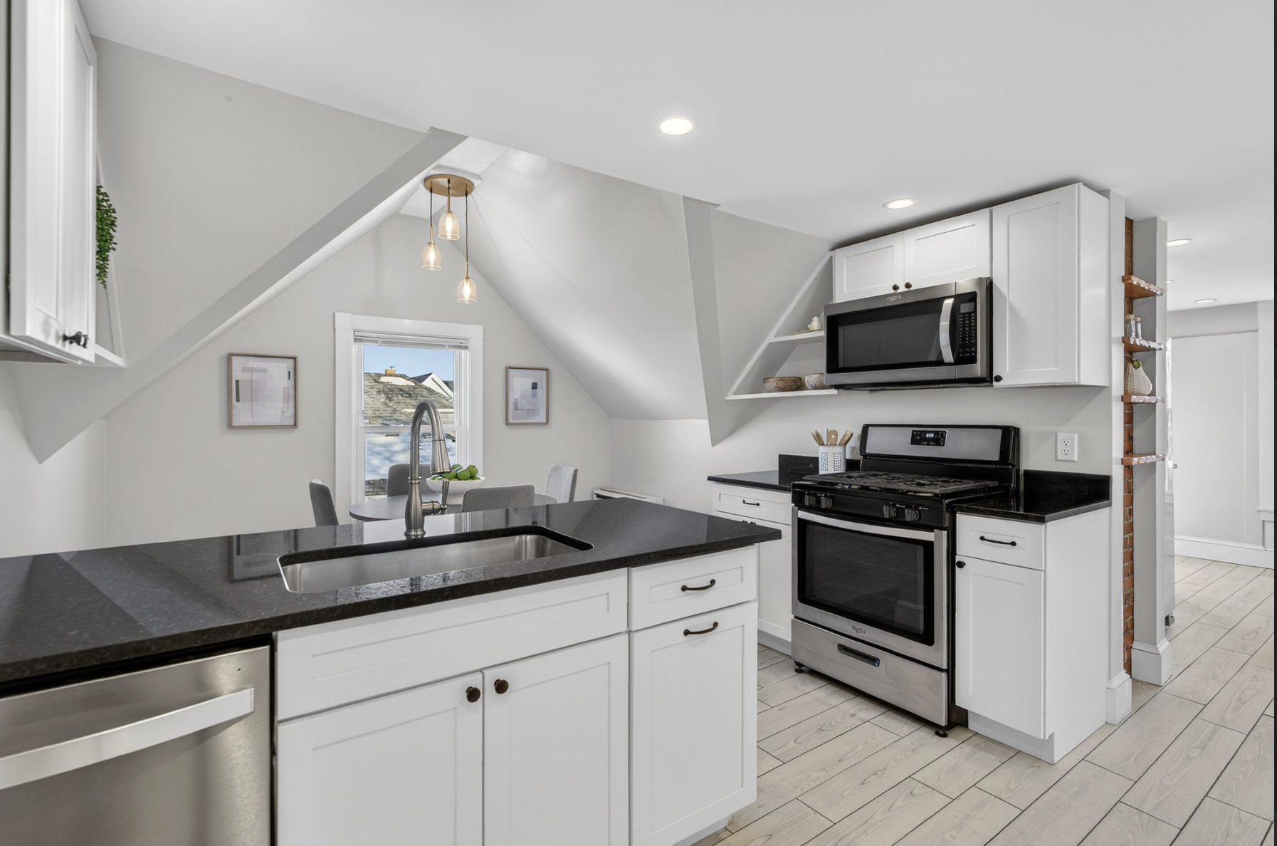Modern kitchen featuring white cabinets, black countertops, stainless steel appliances including a microwave and oven, a kitchen island with a sink, and a window with two framed pictures on the wall nearby.