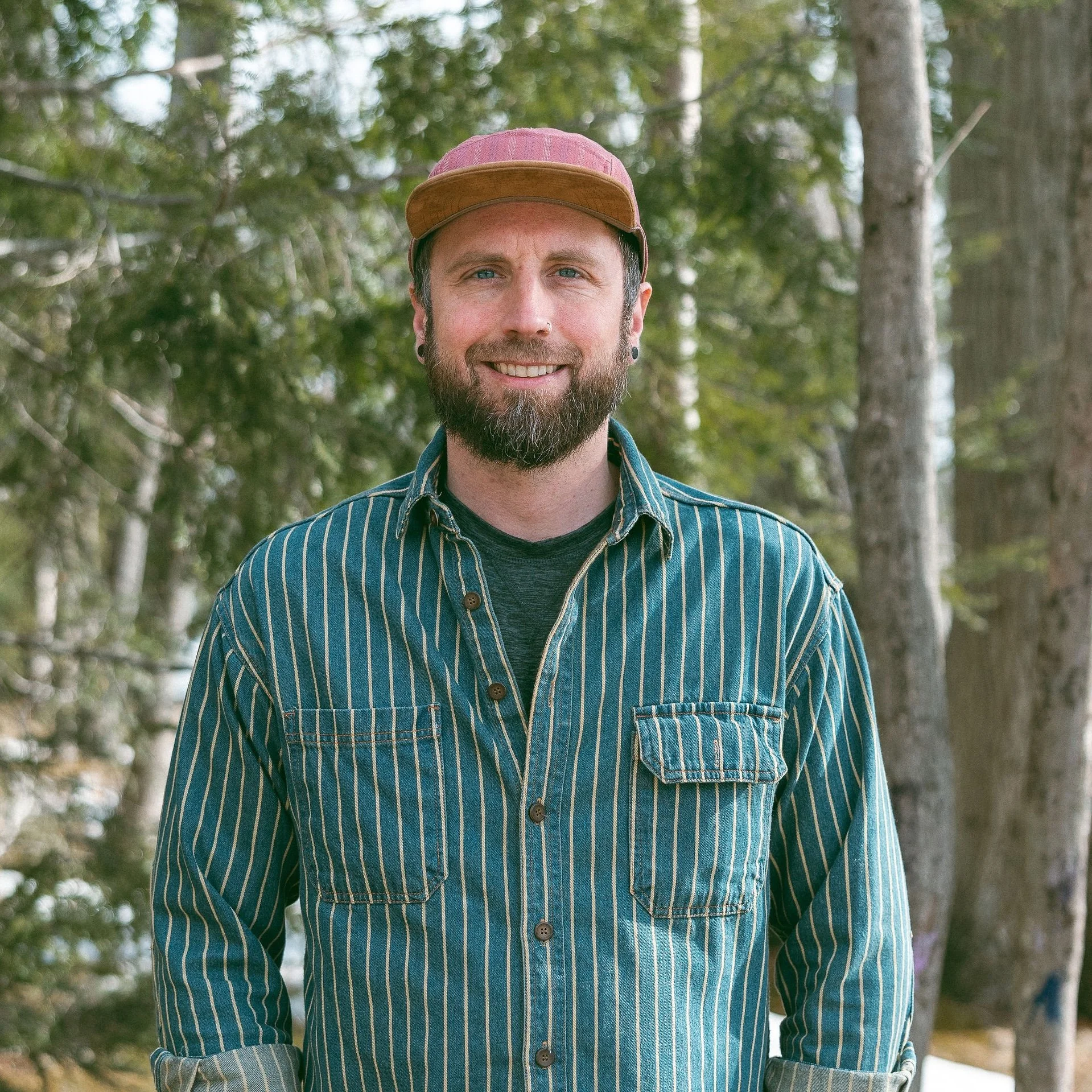 A man with a beard and blue eyes, wearing a striped shirt and a pink and brown cap, smiling outdoors with trees and greenery in the background.