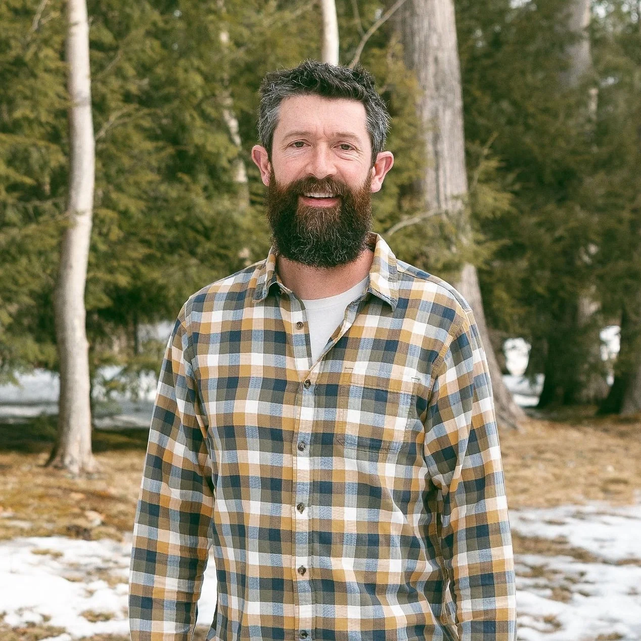 A man with a beard and short dark hair stands outdoors in a forested area with tall trees and patches of snow on the ground, wearing a plaid shirt.