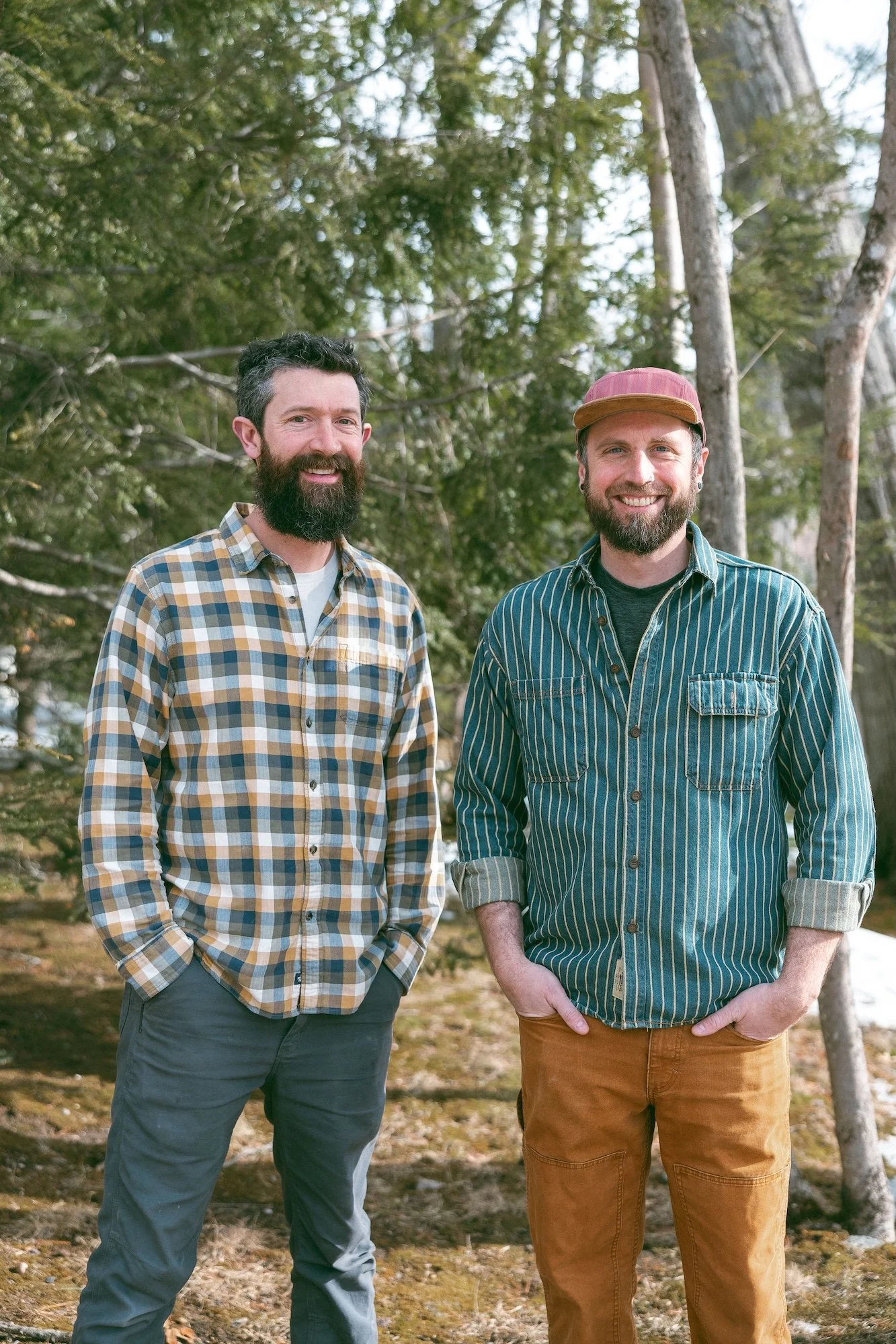 Two men standing outdoors in a wooded area, smiling at the camera. The man on the left has dark hair, a beard, and is wearing a plaid shirt and gray pants. The man on the right has a beard, is wearing a striped shirt, brown pants, and a pink cap.