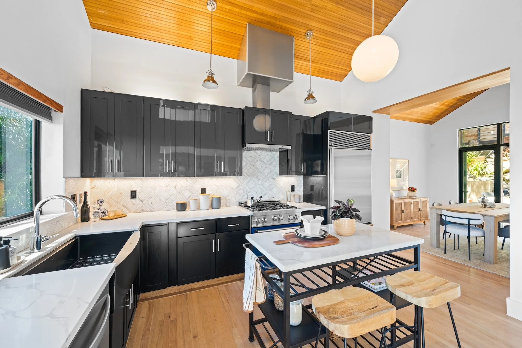 Modern kitchen with black cabinets, white marble countertops, kitchen island with wooden stools, stainless steel appliances, and large windows allowing natural light, with wooden ceiling and a dining area visible in the background.
