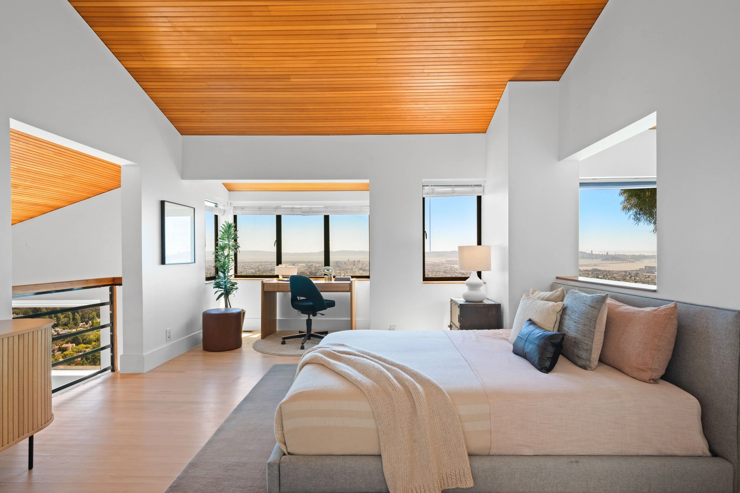 Modern bedroom with hardwood floor, white walls, and large windows offering city and river views. Contains a bed with beige and gray pillows, a nightstand with a white lamp, a desk with a chair, and some indoor plants. Ceiling is covered with wooden panels.