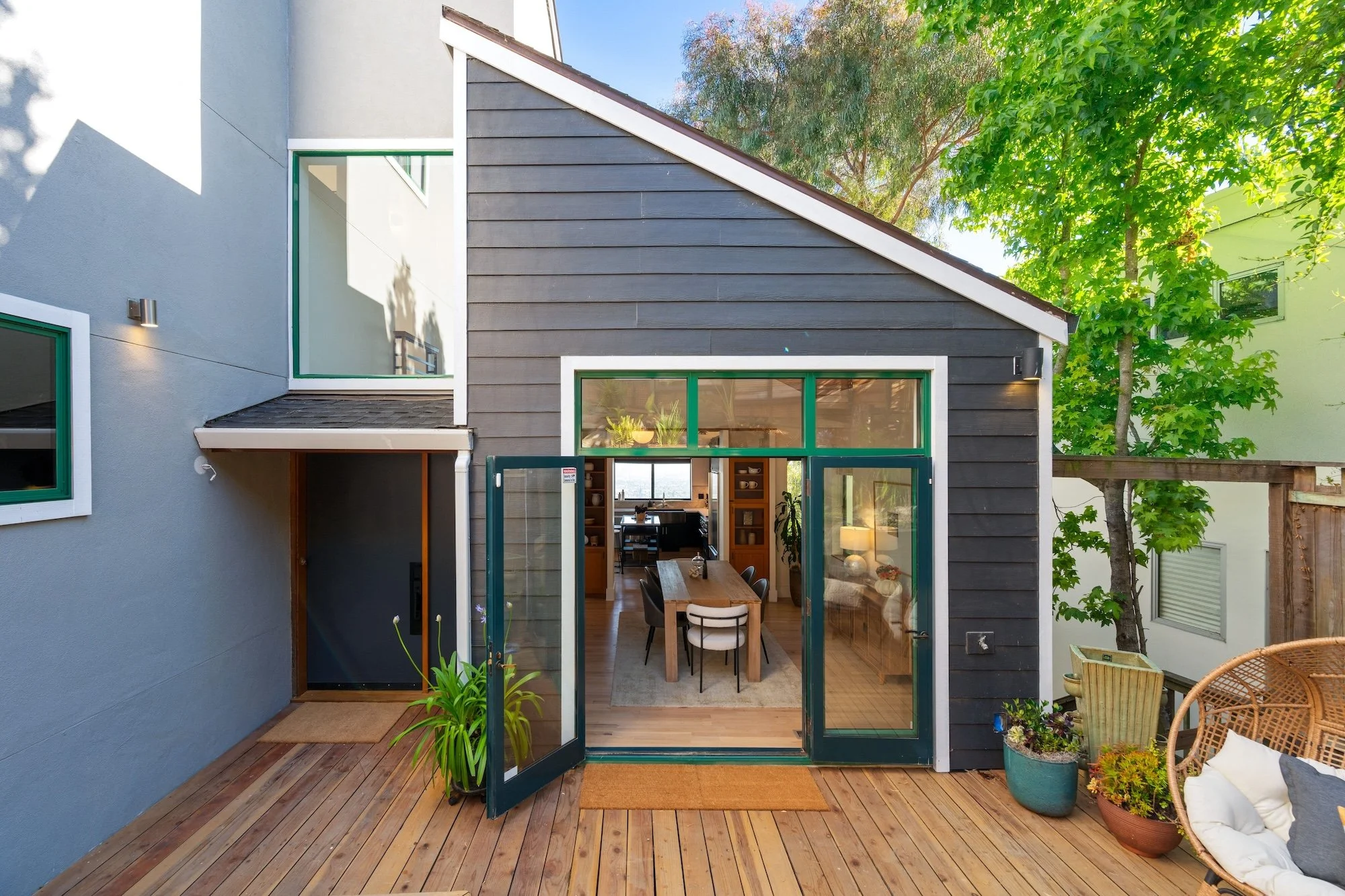 A backyard patio area with wooden flooring, green plants, and large sliding glass doors providing a view into an indoor dining room with a wooden table and chairs. The yard is enclosed by neighboring buildings and has trees with green foliage.