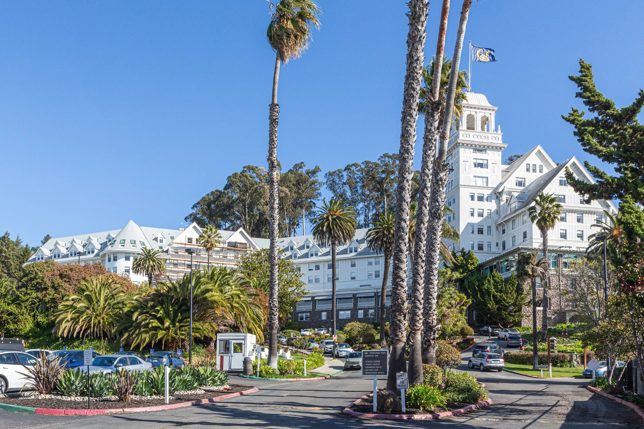A large white hotel with Victorian architecture, multiple gabled roofs, and a tower with flags, surrounded by palm trees and lush greenery. A parking lot with cars is in the foreground.