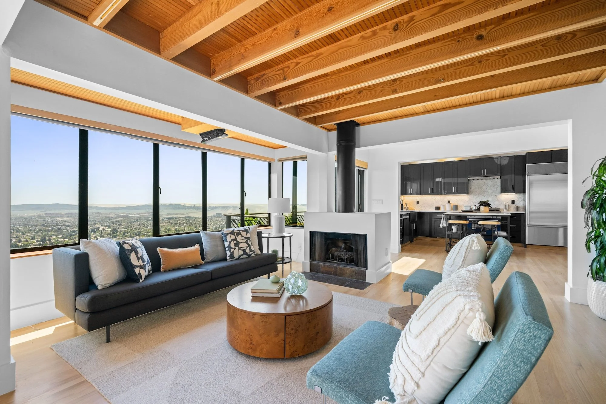 Living room with large windows, a black sofa with patterned and colorful pillows, a wooden coffee table with decorative items, a blue armchair with a shawl, and a white fireplace, with a view of a cityscape and hills outside.