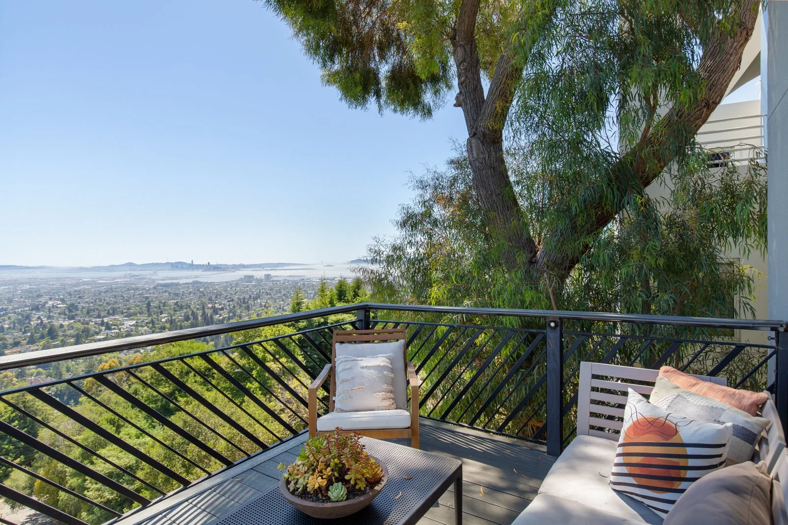 Balcony with outdoor seating area overlooking a cityscape and water in the distance, with a large tree nearby and patio furniture including pillows and a small table with a succulent plant.