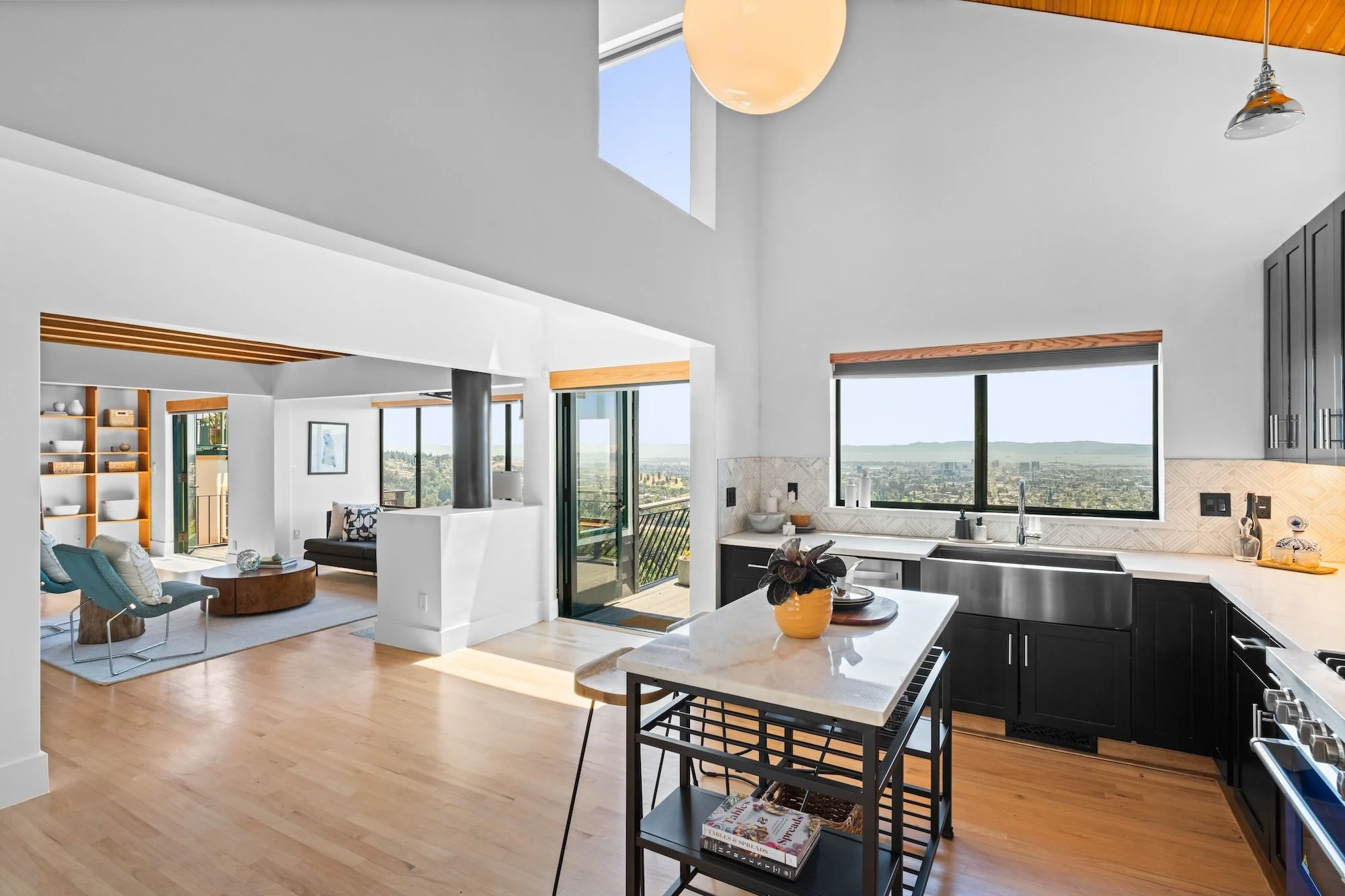 Open-concept kitchen and living room with large windows showing city and landscape views, modern black kitchen cabinets, white potholders, and wooden accents, sunlight streaming in.