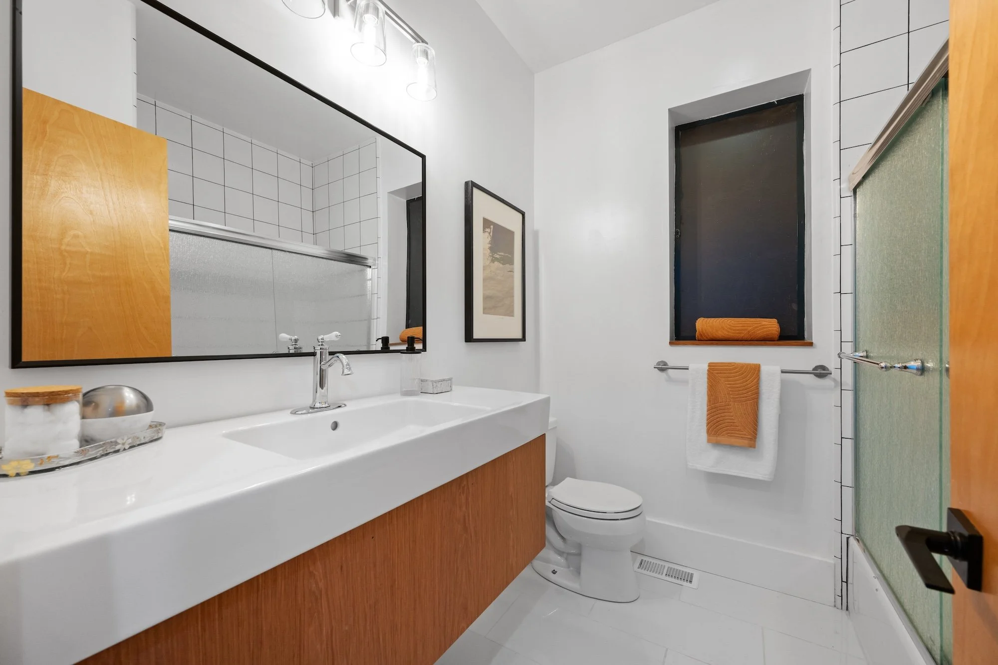 A modern bathroom with a white sink, wooden cabinetry, and a large mirror. The bathroom has black framed windows, white tiled walls, and orange towels. There is a framed picture on the wall and a glass shower enclosure.