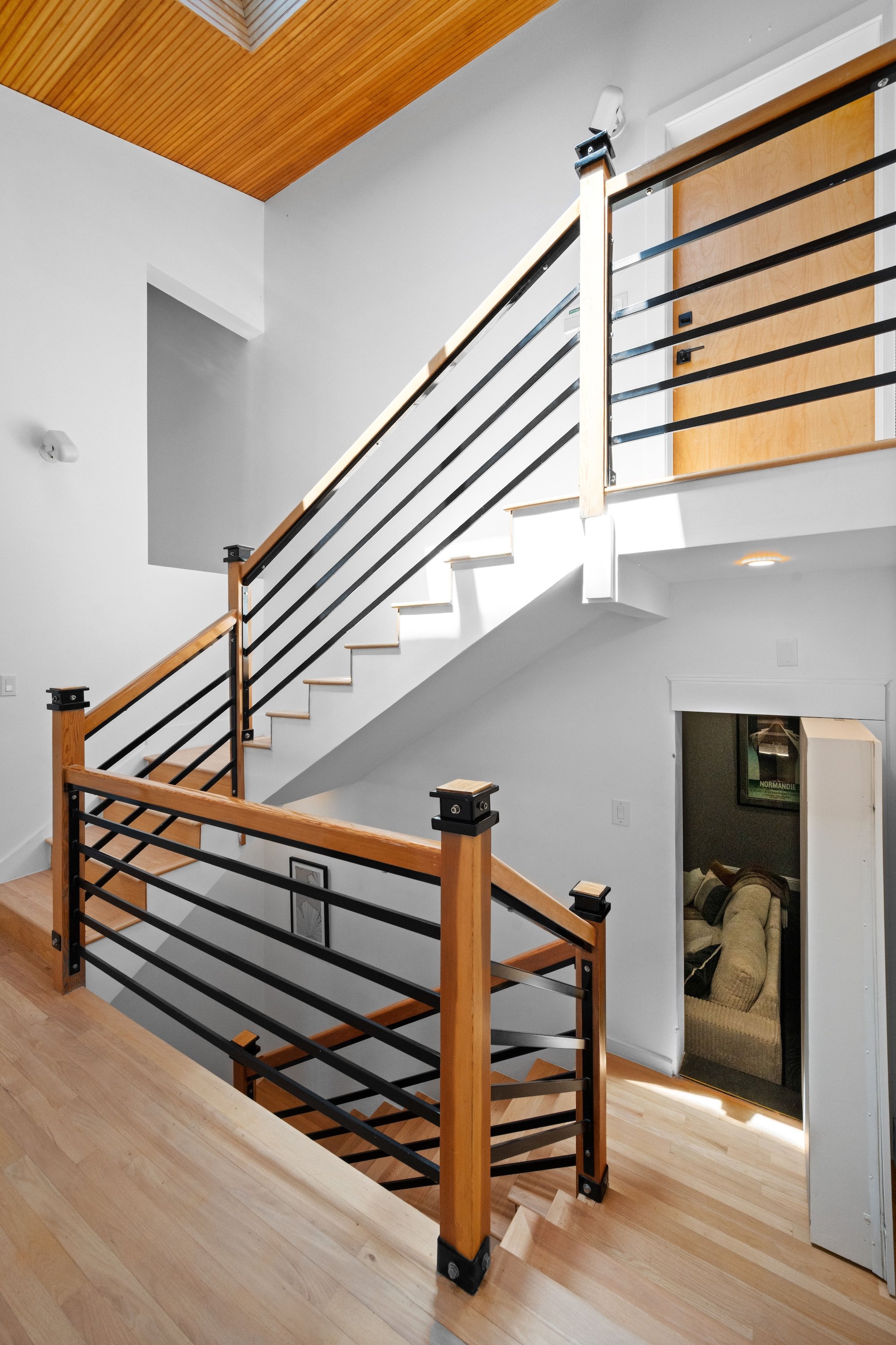 Interior view of a staircase with wooden handrails and black metal balusters, overlooking a living room with a beige couch and a framed poster on the wall.