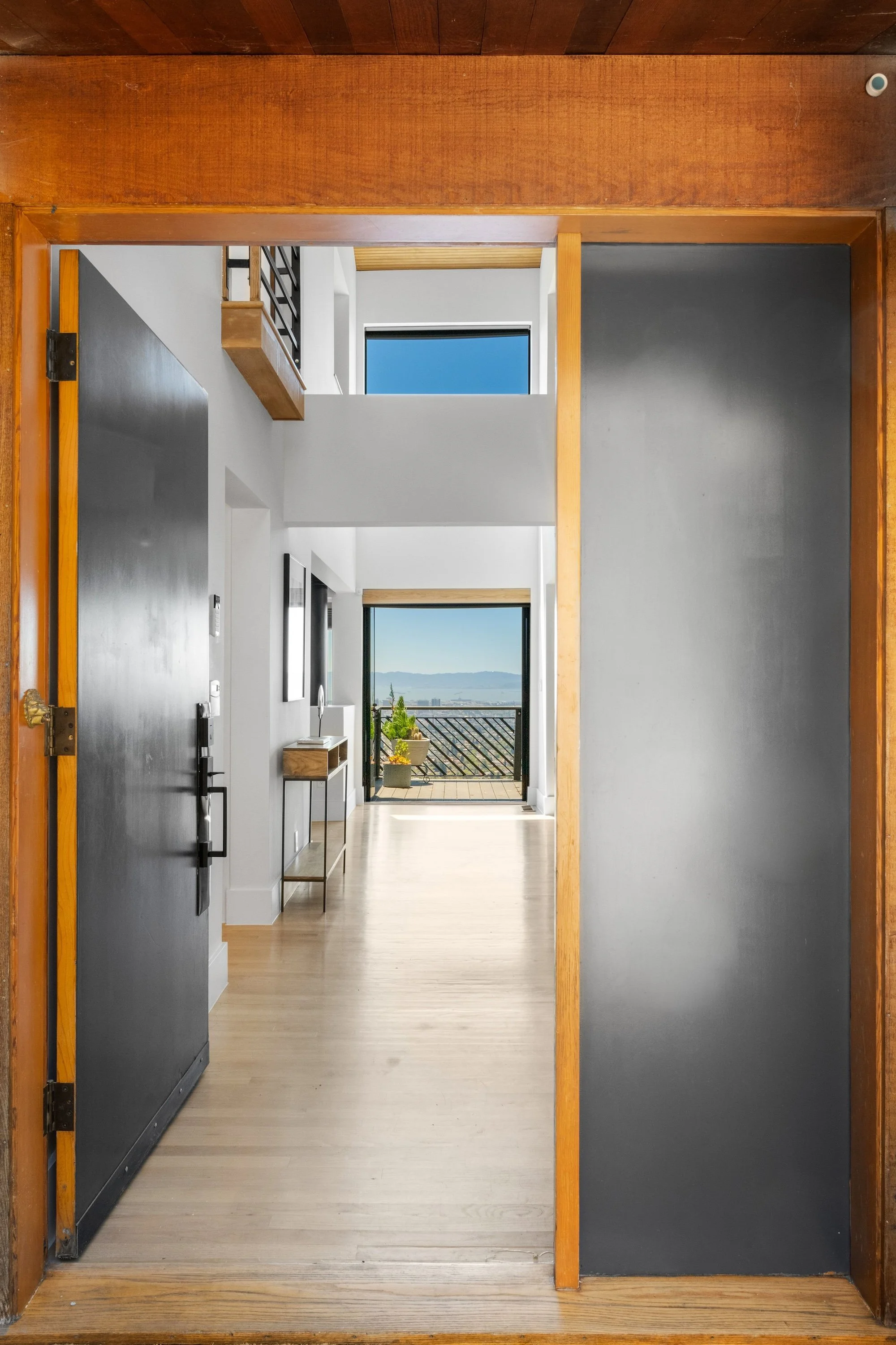 View through an open doorway into a modern apartment hallway with a view of a balcony overlooking a cityscape and water.