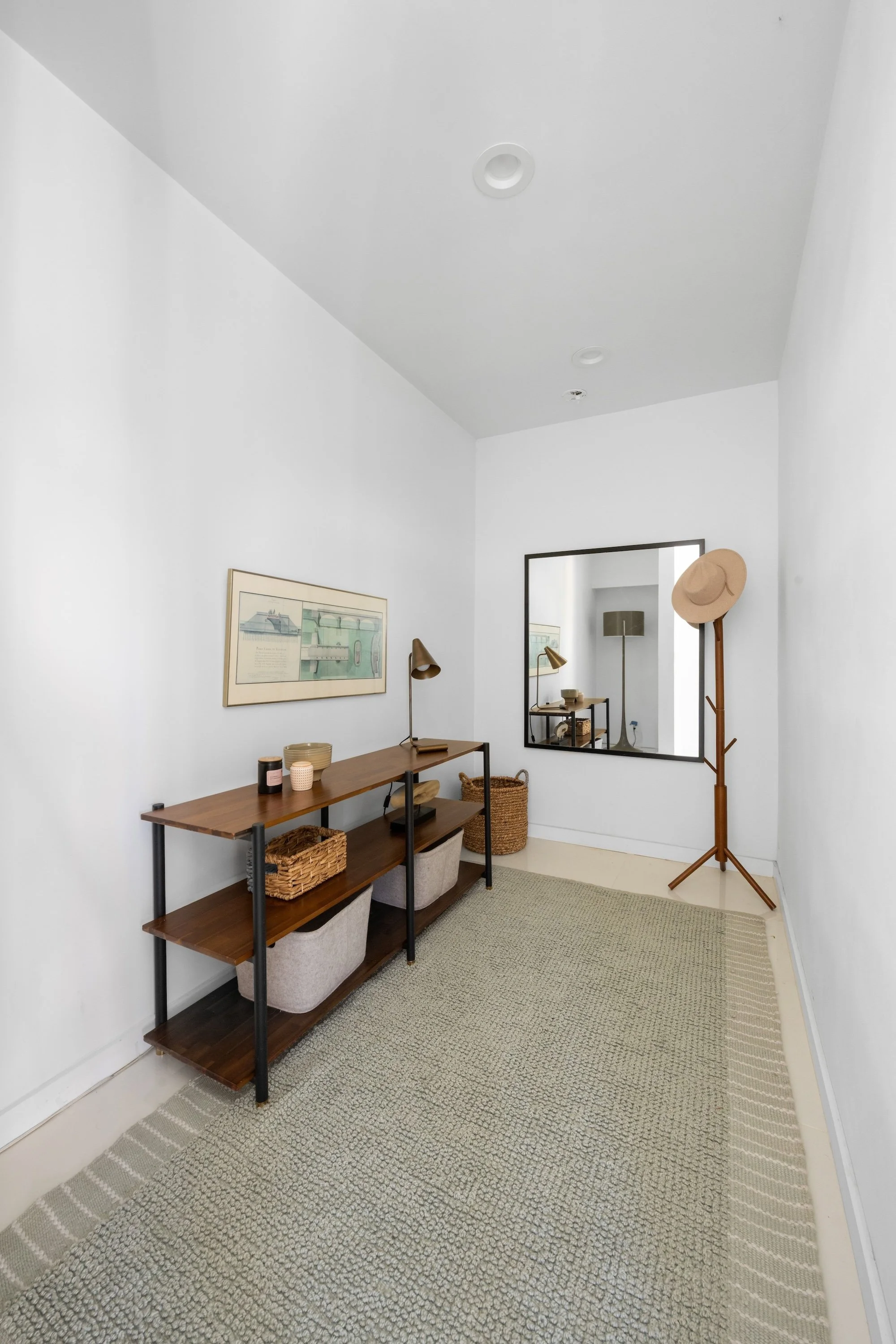 A cozy corner of a room with white walls and minimal decor, featuring a wooden console table with baskets underneath, a wall-mounted mirror, a framed artwork, a brass desk lamp, a standing coat rack with a hat, and a light-colored rug on the floor.