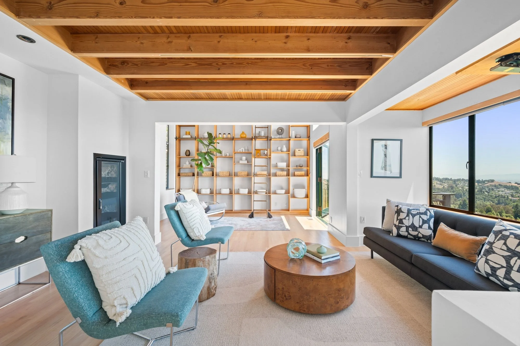 Living room with a high ceiling featuring wooden beams. Contains a blue armchair with a white pillow, a round wooden coffee table with books, a black sofa with patterned pillows, large windows showing a scenic view, a white bookshelf with decorative 