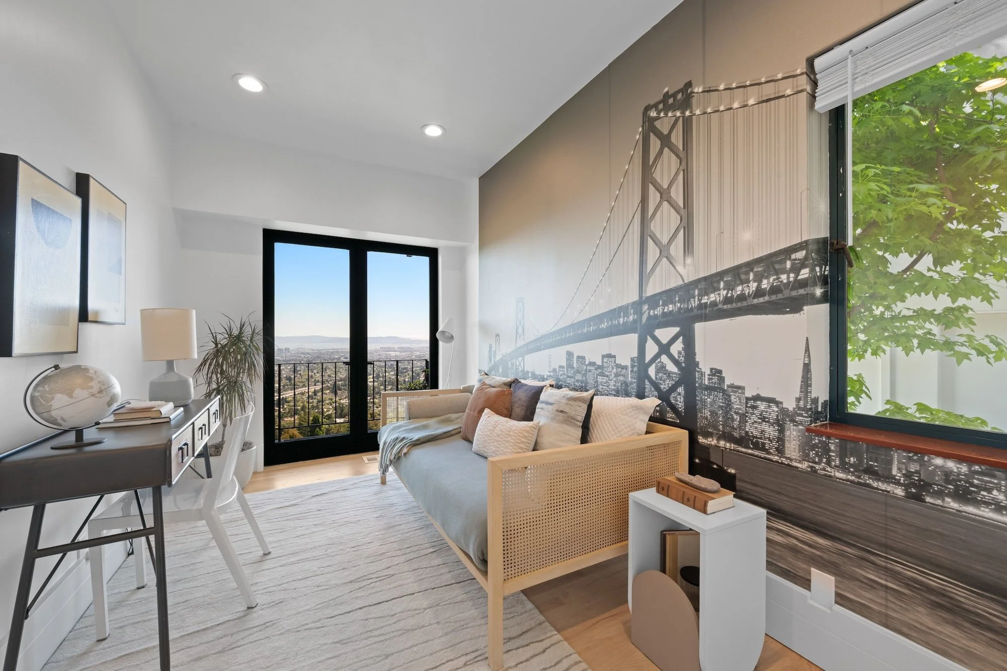 Living room with large bay window, city view, and a wall mural of a bridge and city skyline, featuring a beige sofa, white side table, potted plant, and artwork on the wall.