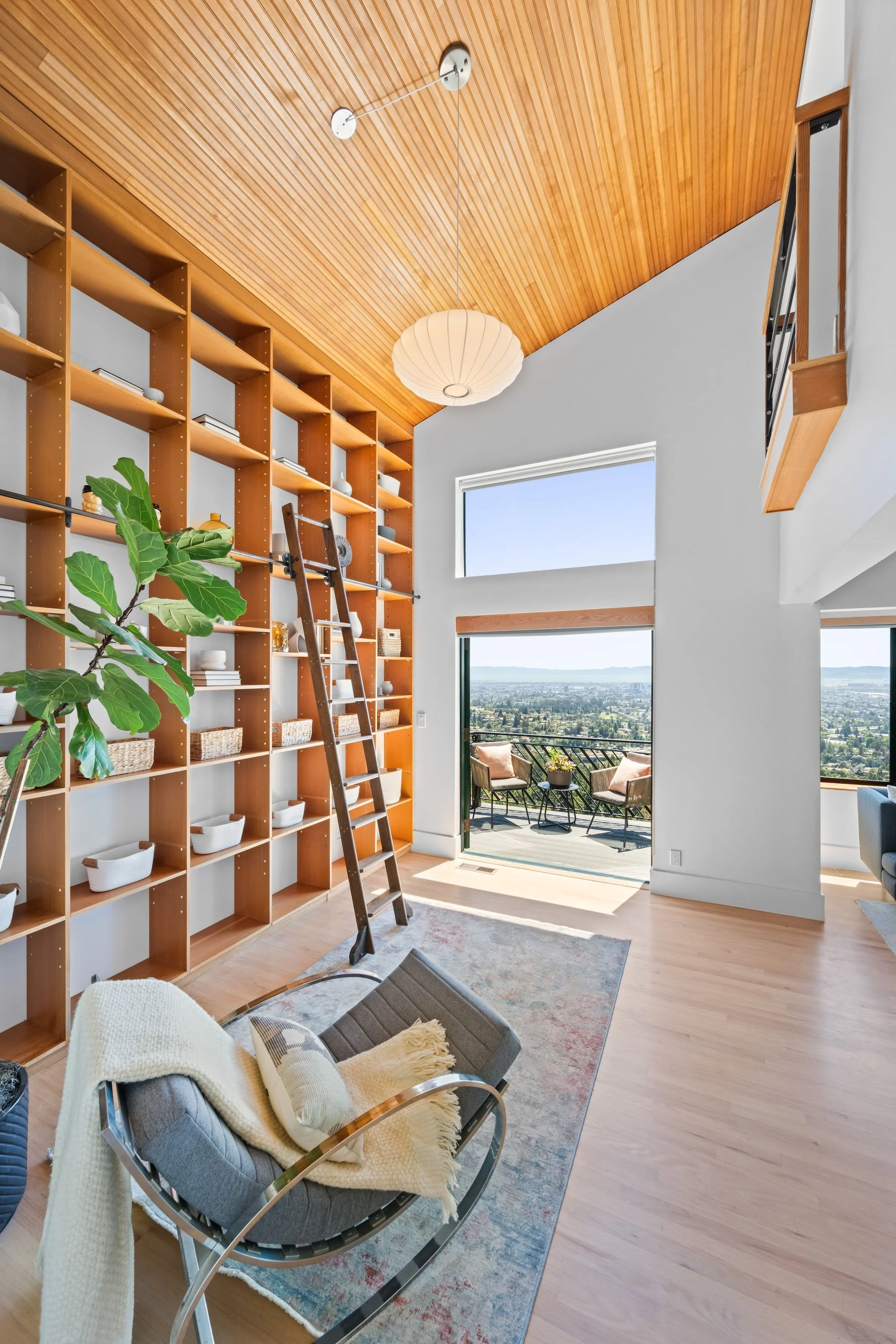 Living room with wooden vaulted ceiling, large shelving unit, ladder, and a balcony with outdoor chairs and a view.