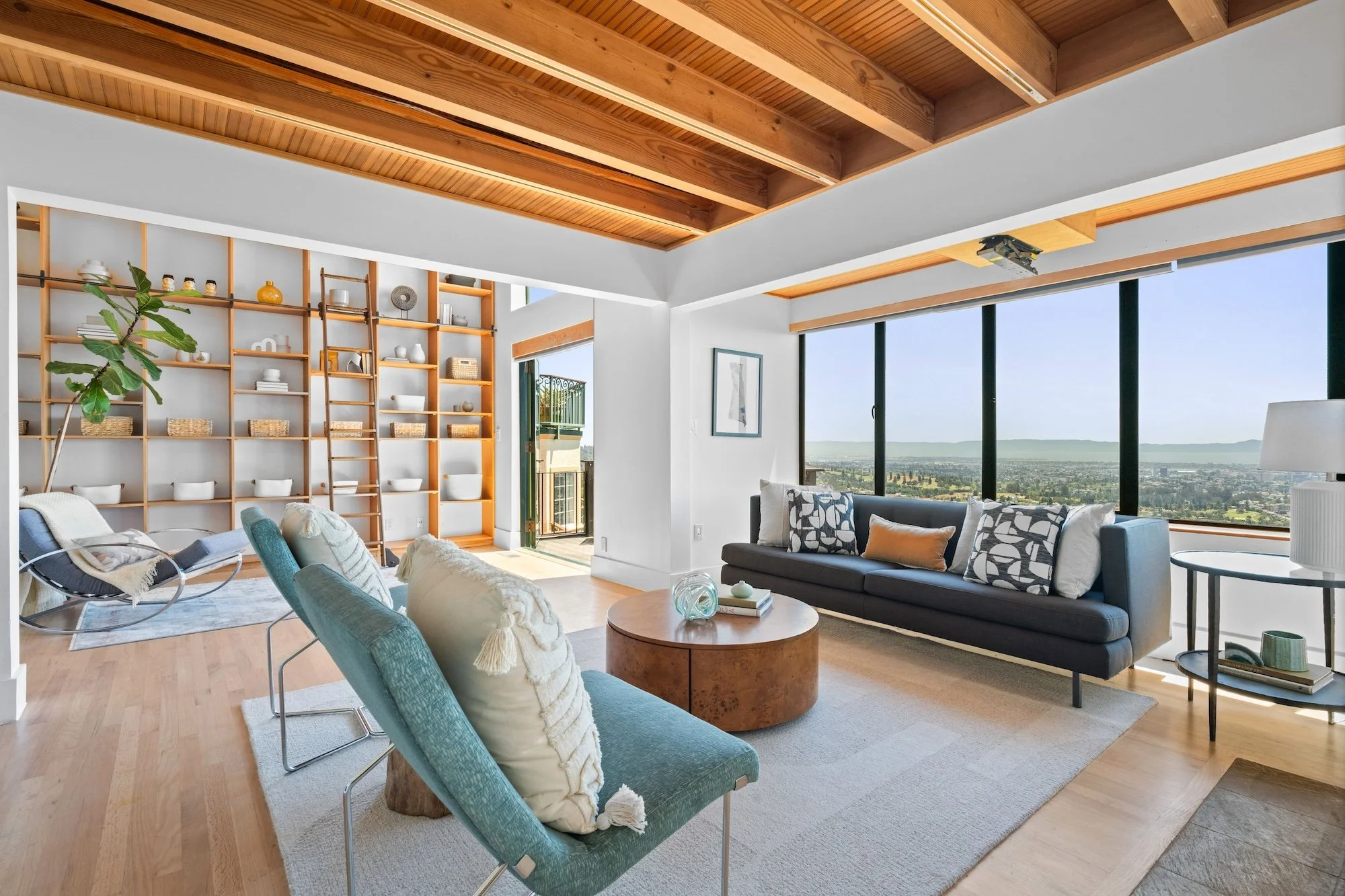 Modern living room with large windows, wooden ceiling, white walls, and a mix of colorful seating including blue, gray, and beige chairs and sofas. A tall bookshelf with decorative items and baskets, a small balcony visible through the open door, and