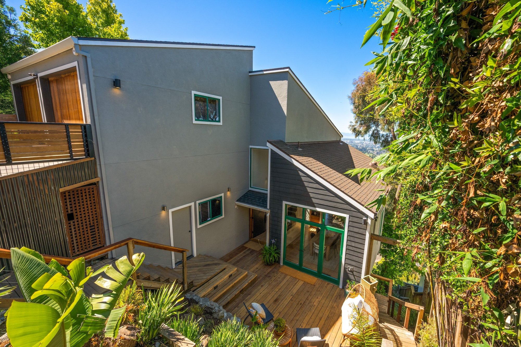 Modern residential building with a gray exterior, multiple windows, a small balcony with a wooden railing, and a backyard with wooden decking and lush green plants, set against a bright blue sky.
