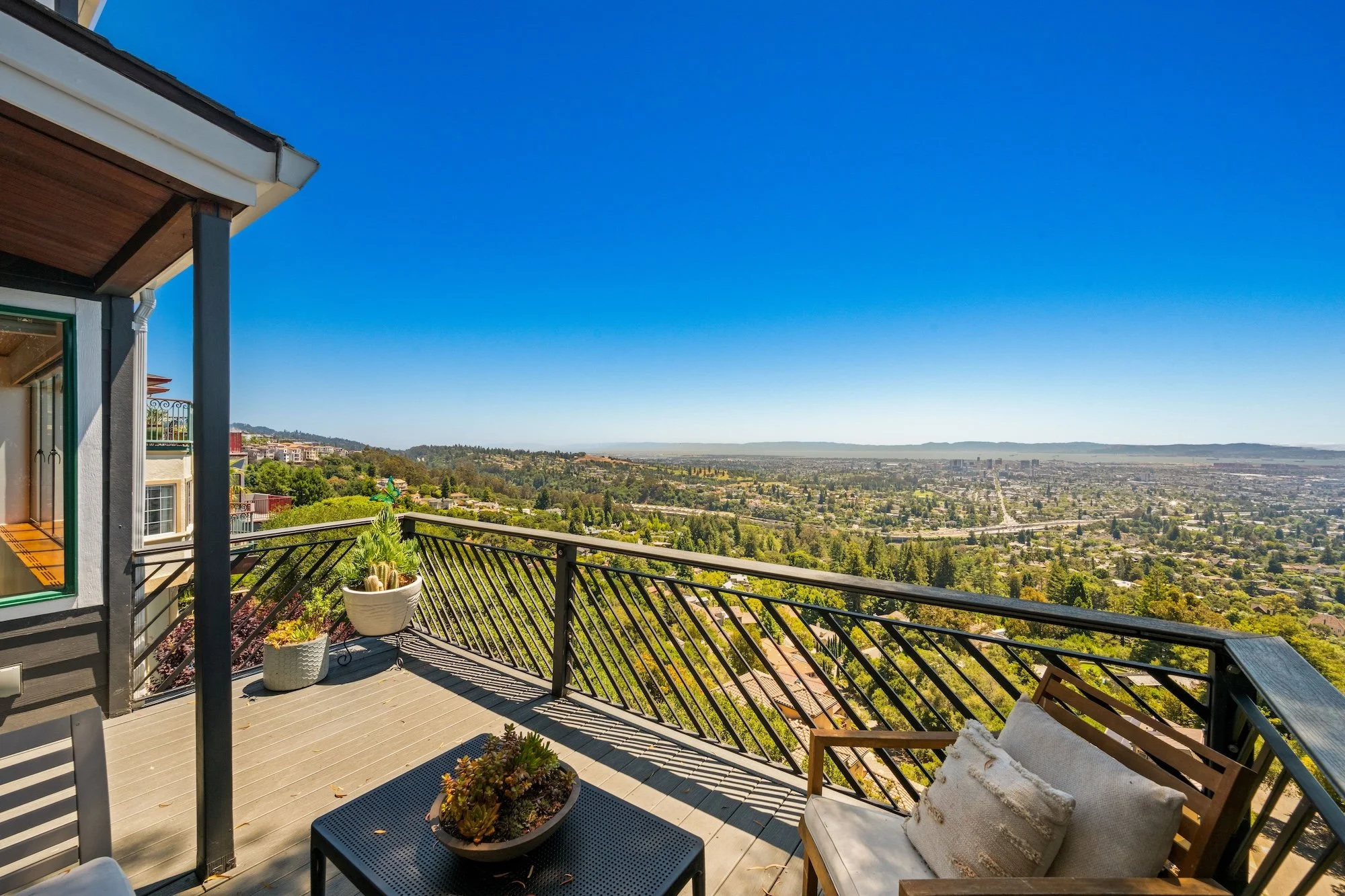 A spacious balcony with outdoor furniture including a black table with potted plants, a wooden chair with a cushion, and a seating area with pillows overlooking a city and green hills under a clear blue sky.