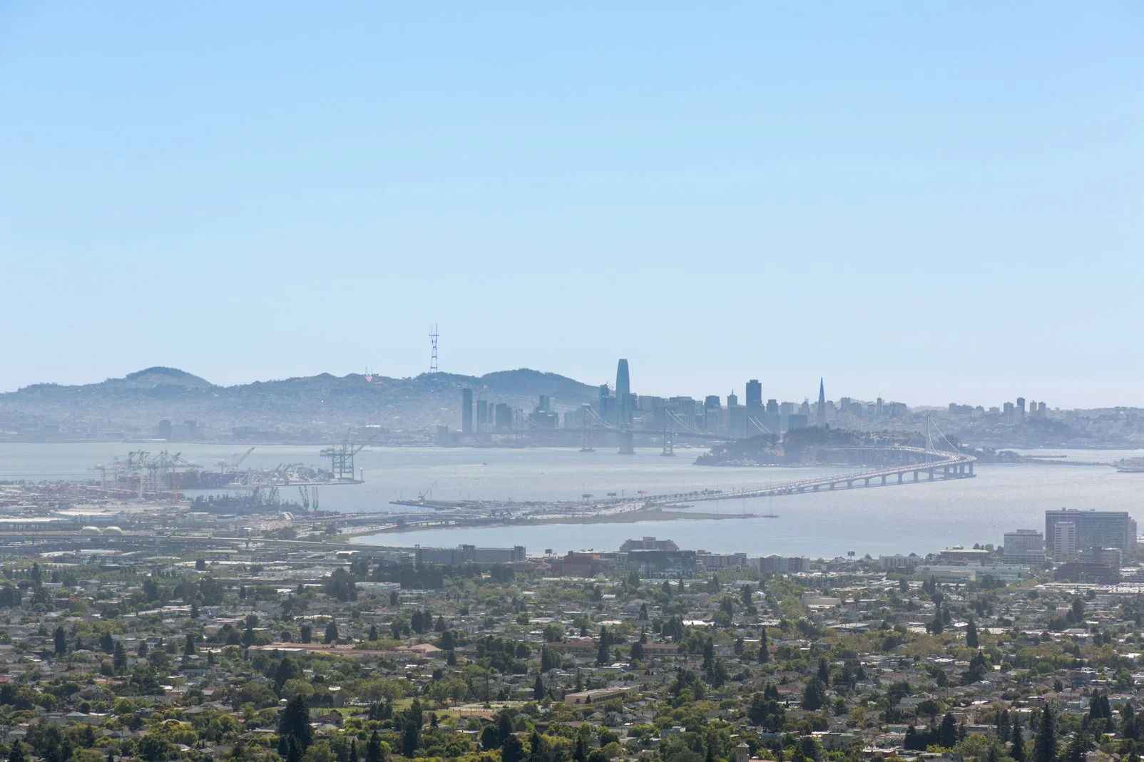 A city skyline with tall buildings, a bridge, and hills in the background, viewed across a body of water with a cityscape in the foreground.