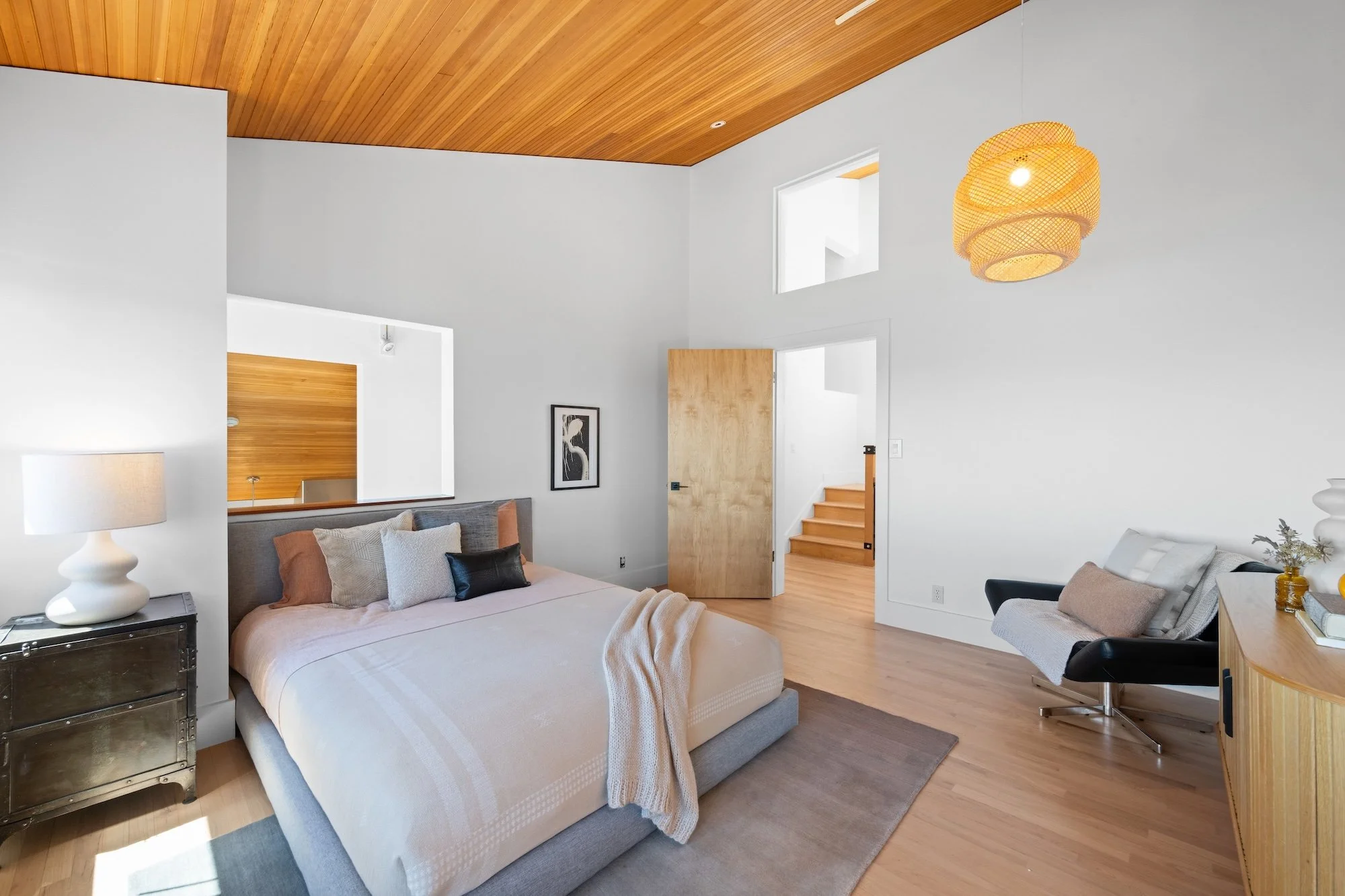 Modern bedroom with elevated ceiling, wooden paneling, and minimalistic decor. Features a large bed with beige and brown pillows, a bedside table with a white lamp, and a sitting area with a black chair. There is a wooden door, a staircase, and small