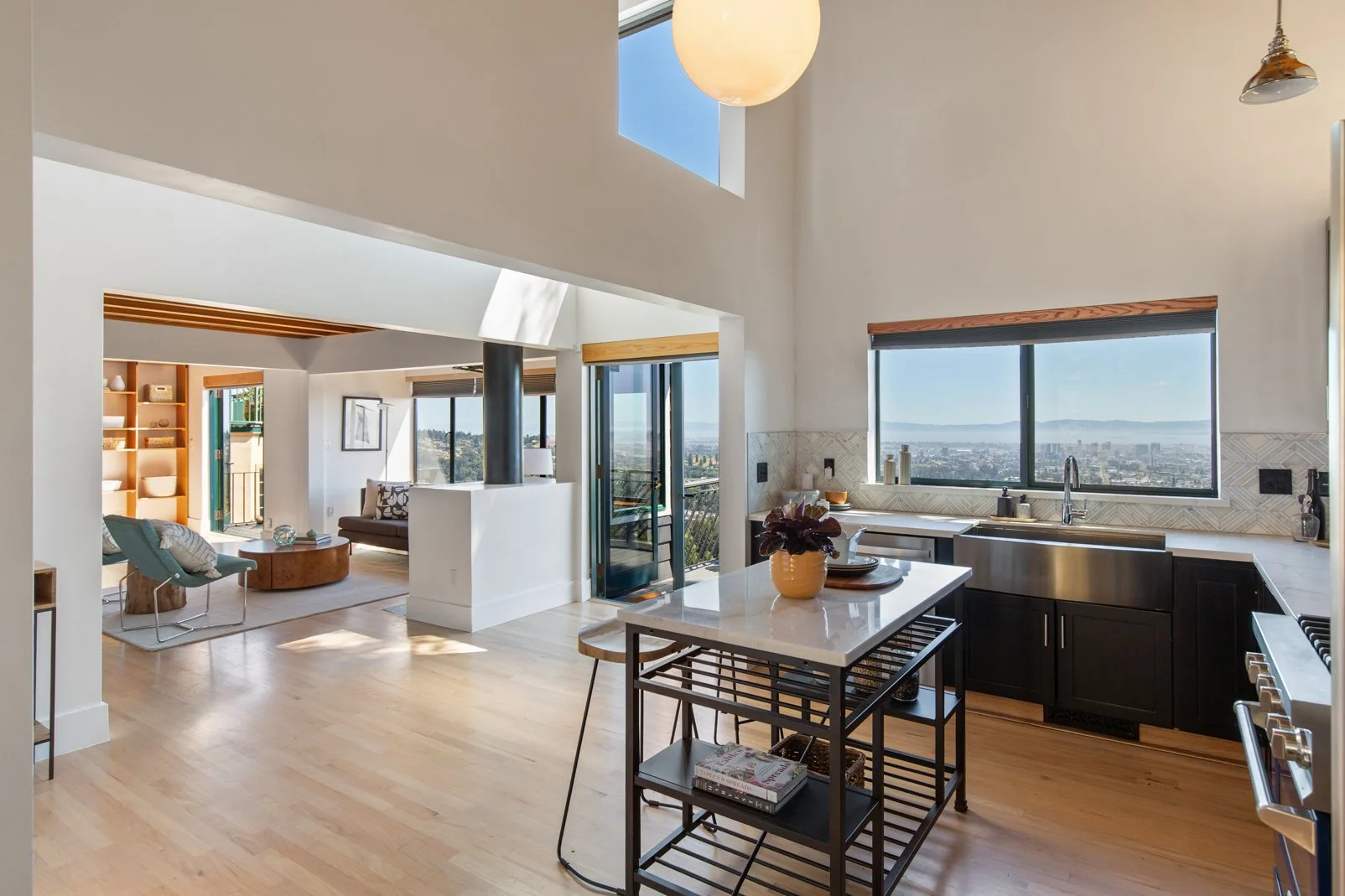 Open-concept modern kitchen and living room with large windows, hardwood floors, and minimalist decor, overlooking a cityscape and water.