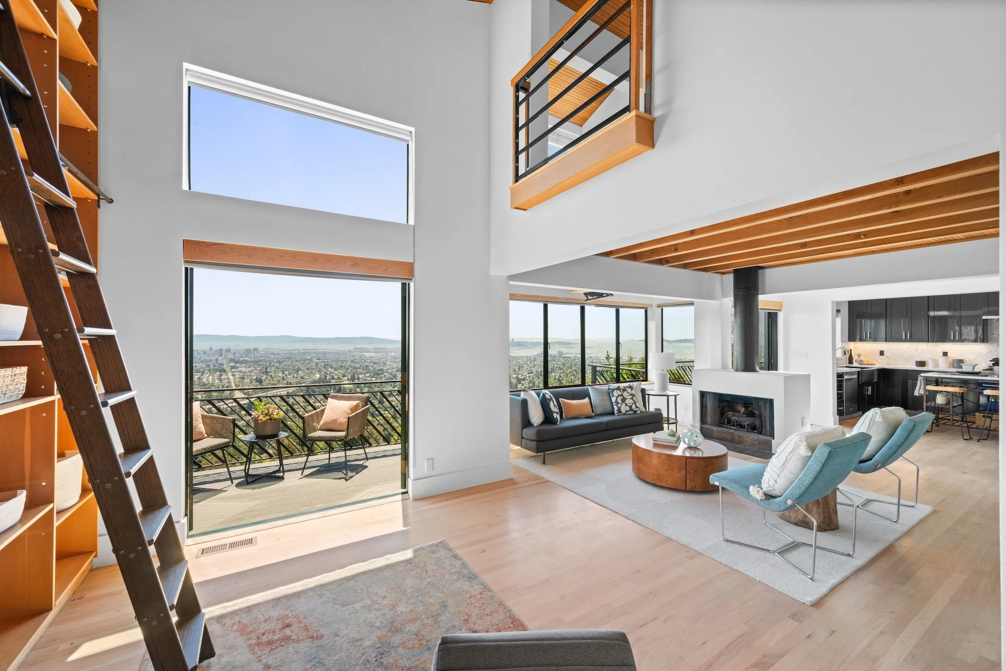 A spacious living room with large windows and a balcony that offers city and mountain views. The room has modern furniture, including a black sofa, a light blue armchair, and a round wooden coffee table. There is a fireplace with a black chimney, and
