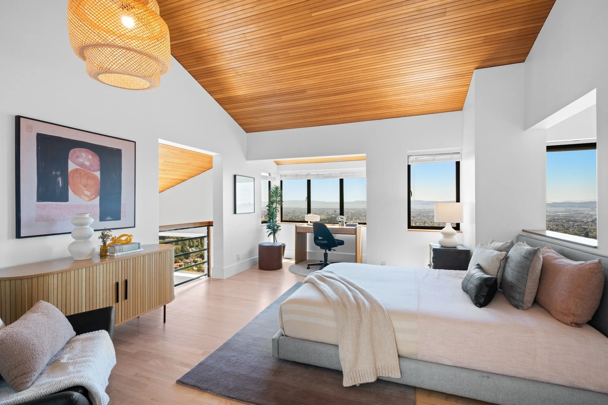 Modern bedroom with large windows showing city view, white walls, wooden ceiling, with a bed, sofa, art on wall, and decorative items.