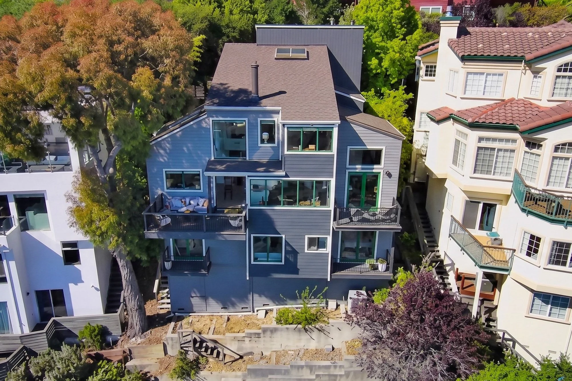 An aerial view of a multi-story modern house with gray siding, large windows, and outdoor balconies. The house is surrounded by tall trees and neighboring houses with tiled roofs.