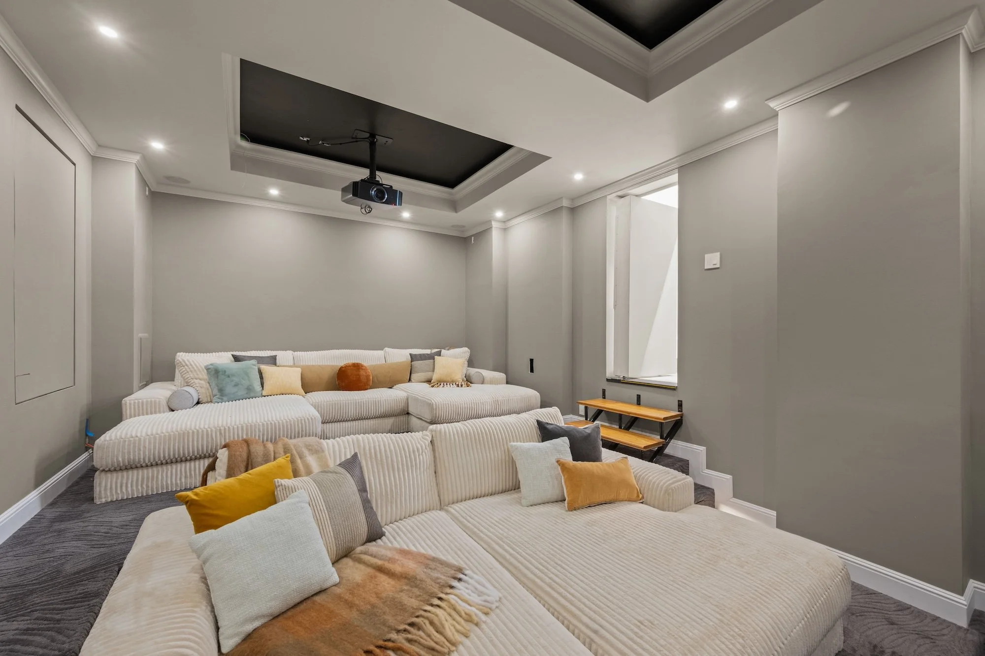 A modern living room with gray walls and white crown molding. There is a large beige sectional sofa decorated with various pillows, including yellow, gray, and patterned ones. A small wooden step stool is next to the sofa, and a black ceiling-mounted