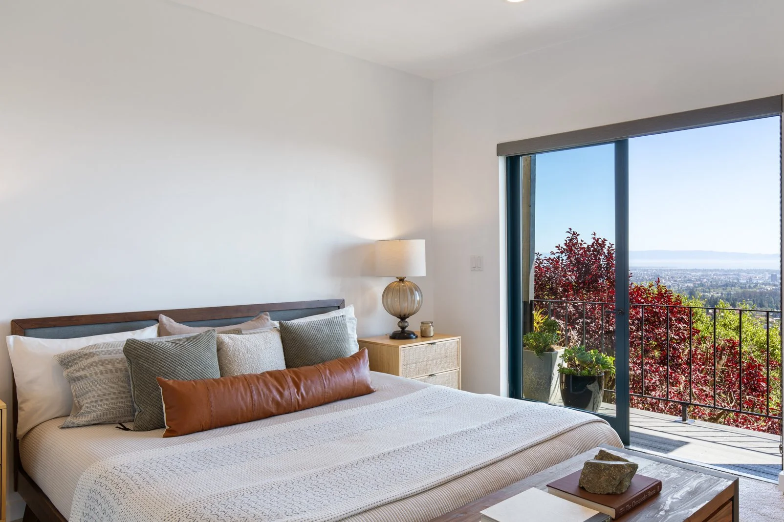 Bedroom with a bed, pillows and a nightstand with a lamp, open sliding door leading to a balcony with potted plants, trees, and a city view in the distance.