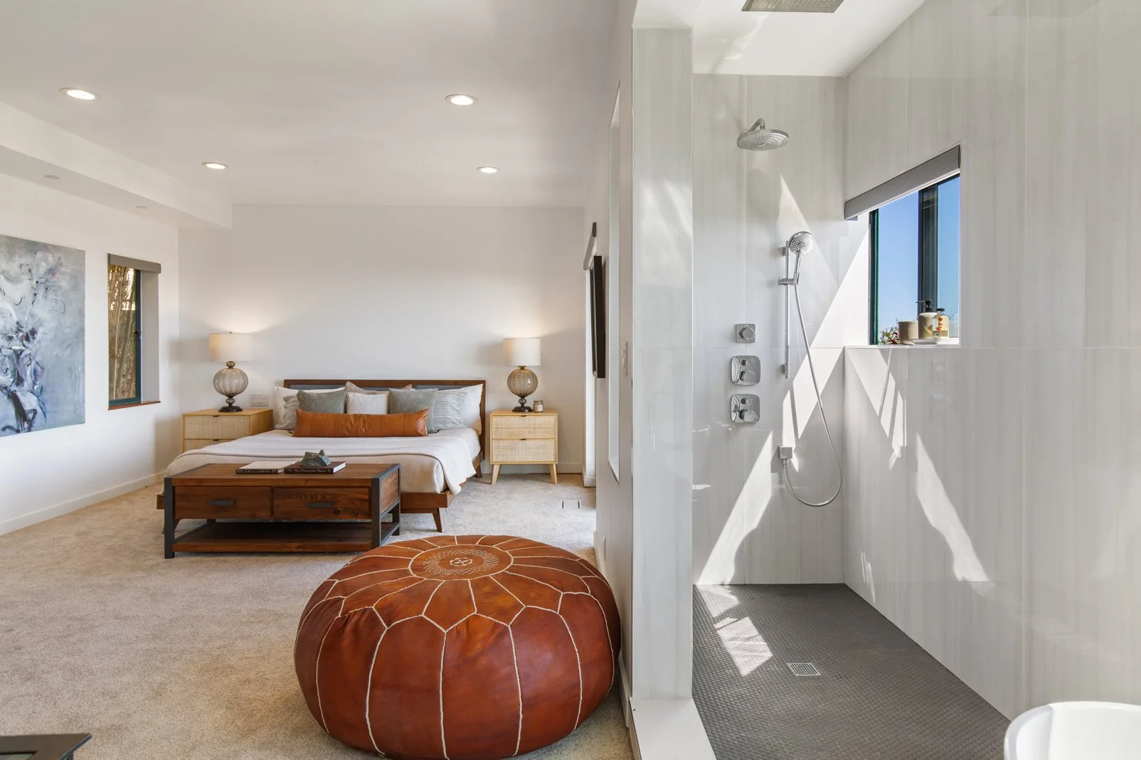 A bedroom with a bed, nightstands, lamps, and a painting on the wall, with an open bathroom shower area visible on the right side.