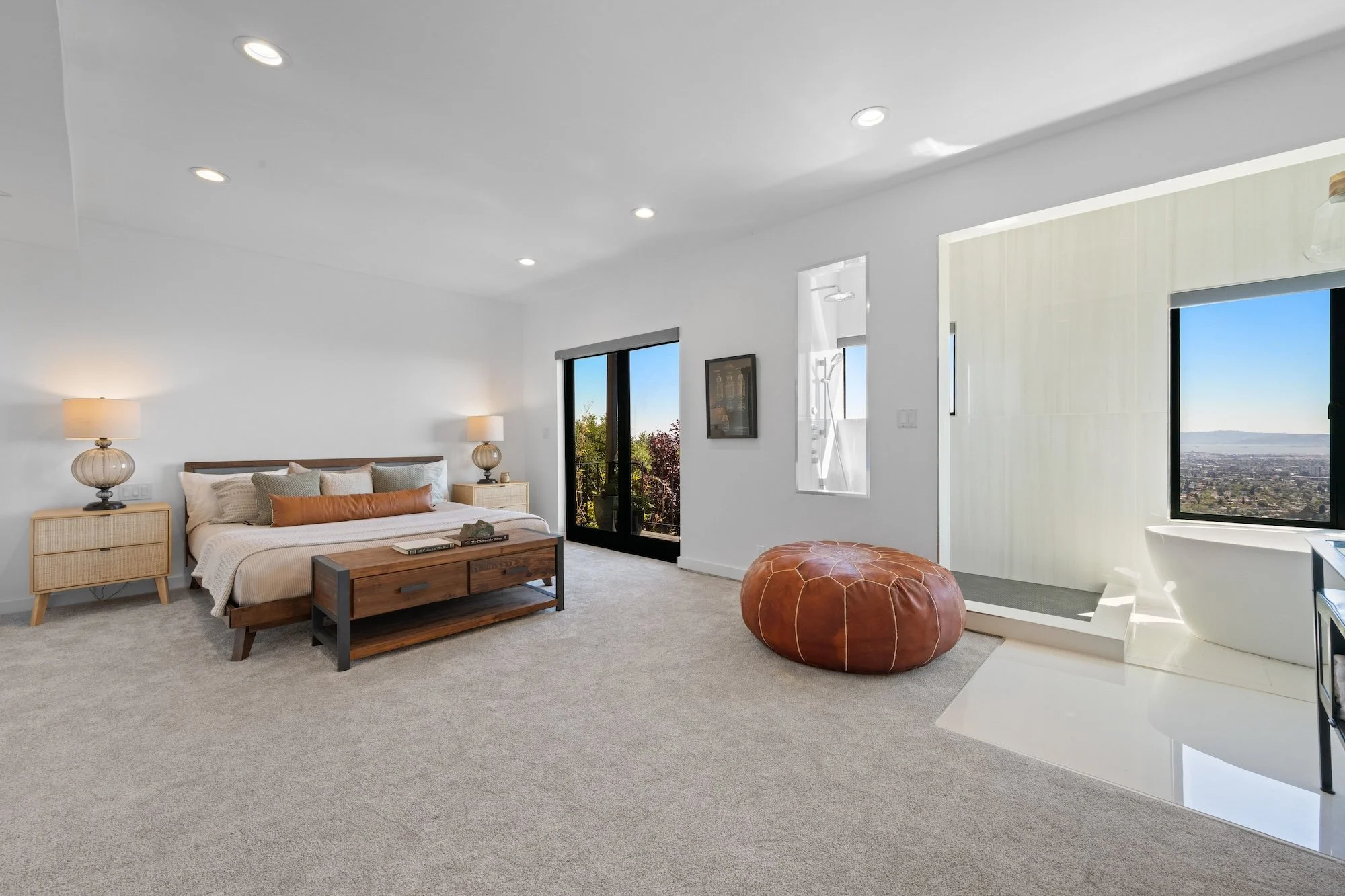 Bright modern bedroom with large window and balcony door, minimal decor, beige carpet, bed with beige and brown pillows, two nightstands with lamps, leather pouf, and mountain view.