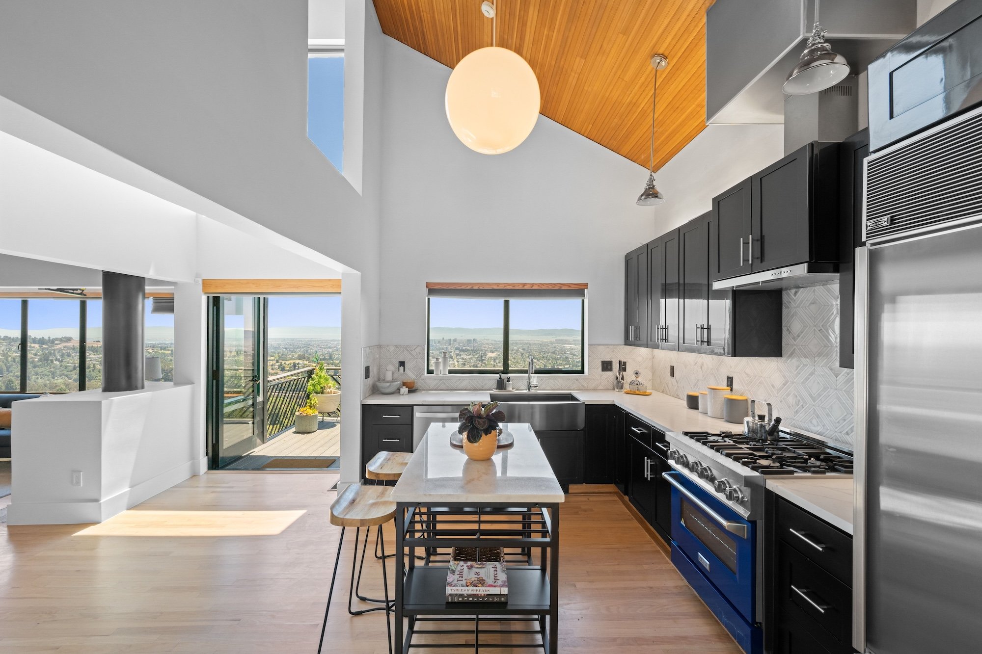 Modern kitchen with black cabinets, stainless steel appliances, wooden flooring, and a sloped wooden ceiling. Large windows and a glass door lead to an outdoor balcony with city views. A white island with a potted plant is in the center, and a table 