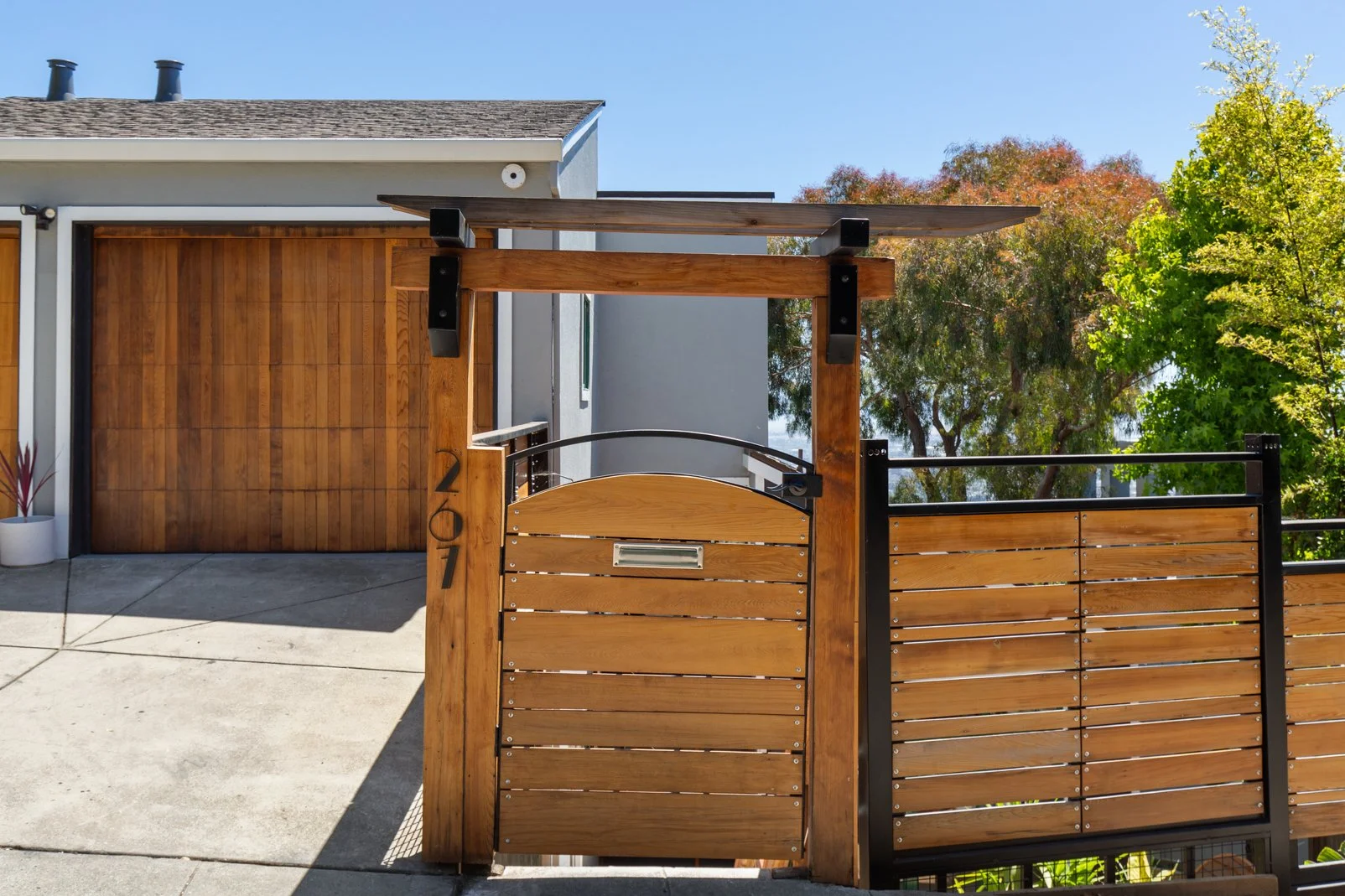Modern house entrance with wooden gate, mailbox, and wooden garage door, surrounded by trees and a concrete driveway.