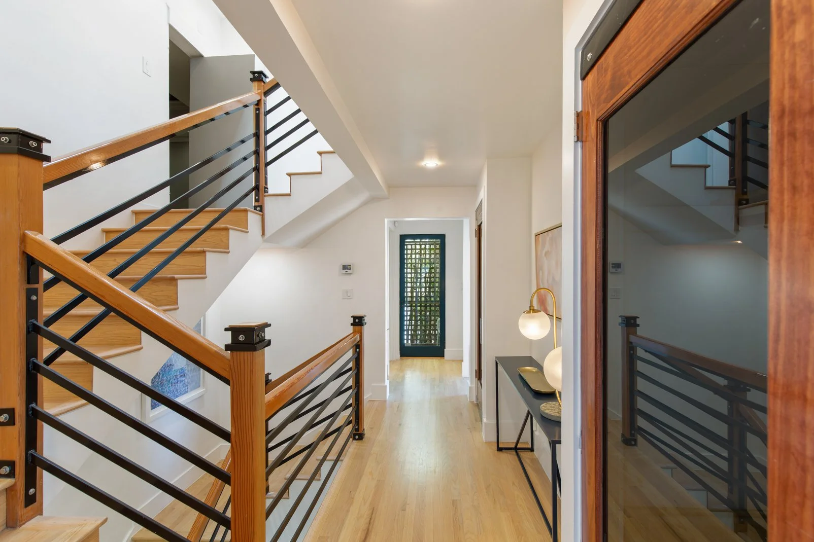 Interior view of a modern house entryway with wooden staircase, black metal railings, a door with a grid window at the end, and a small console table with a lamp.