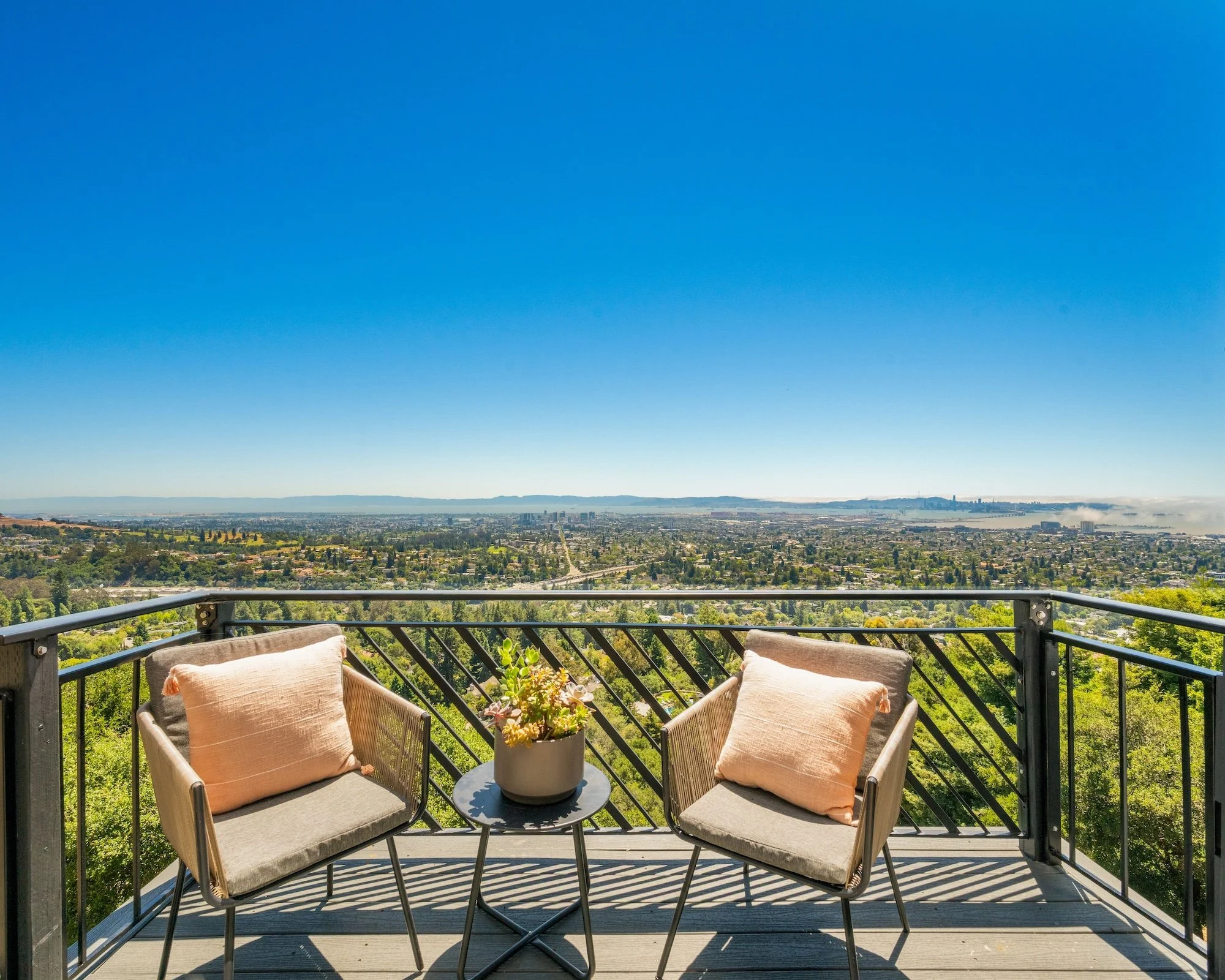 A balcony with two outdoor chairs and a small round table holding a potted succulent plant, overlooking a sprawling cityscape with green areas, distant buildings, and a body of water under a clear blue sky.