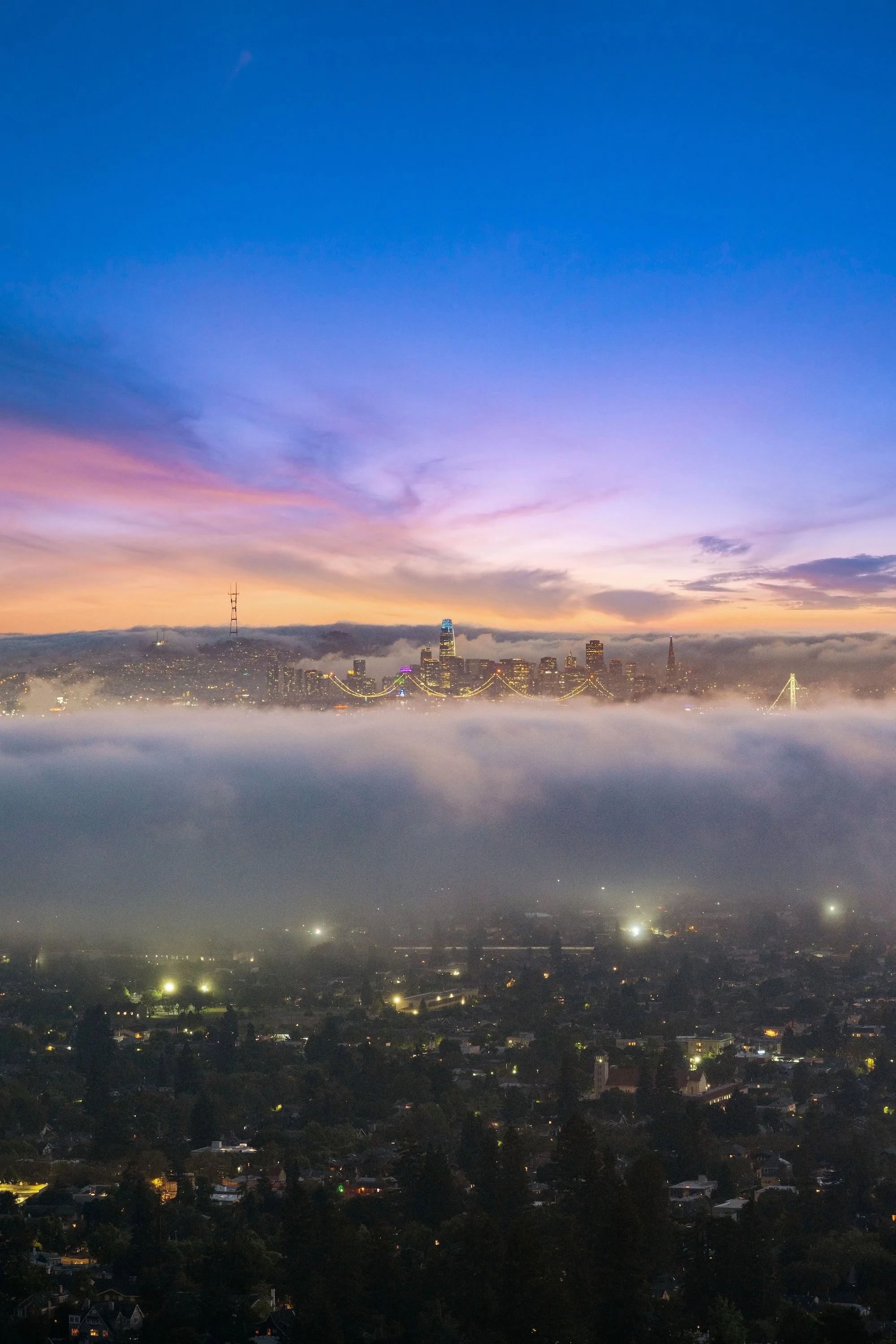 A city skyline at dawn with a colorful sky, fog covering the city, and illuminated buildings and bridges