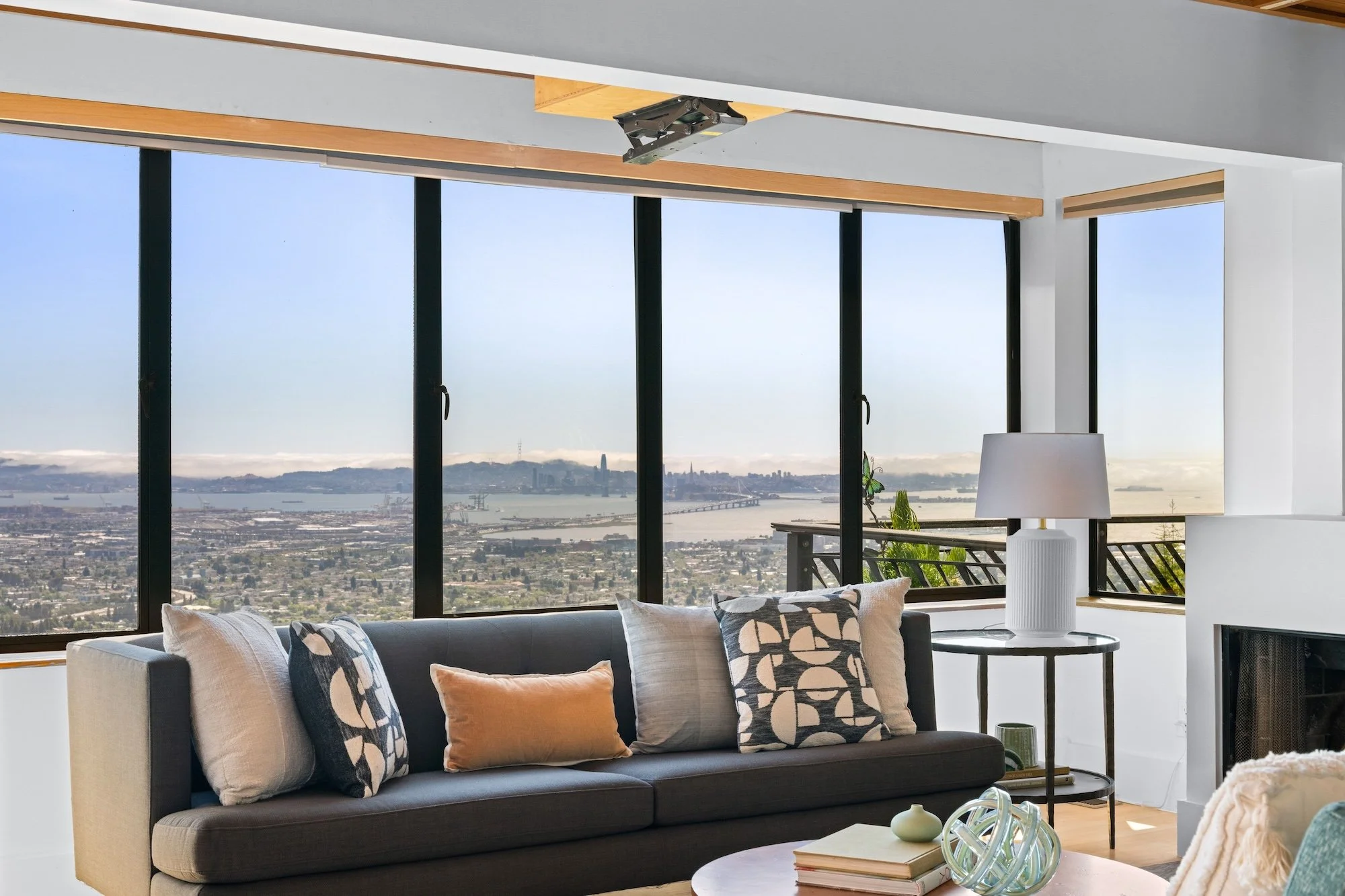 Living room with large windows revealing a cityscape and water view. The room has a dark sofa with patterned and plain throw pillows, a round side table with decorative items, a white lamp with a textured base, and a bookshelf.