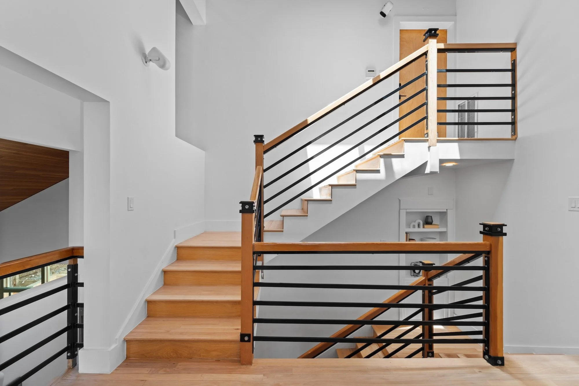 Interior view of a modern staircase with wooden steps, metal and wood railing, white walls, and hardwood flooring.