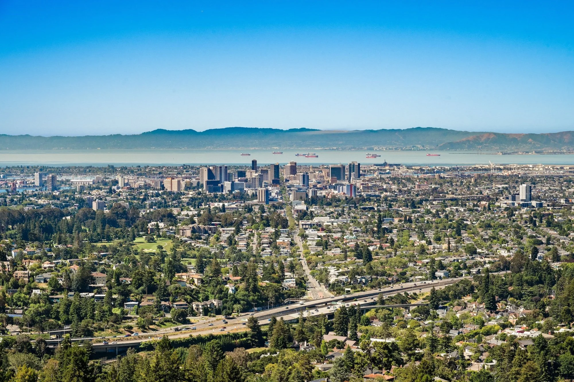 A city skyline with tall buildings near a coastline and ships in the water, with mountains in the background and a clear blue sky.