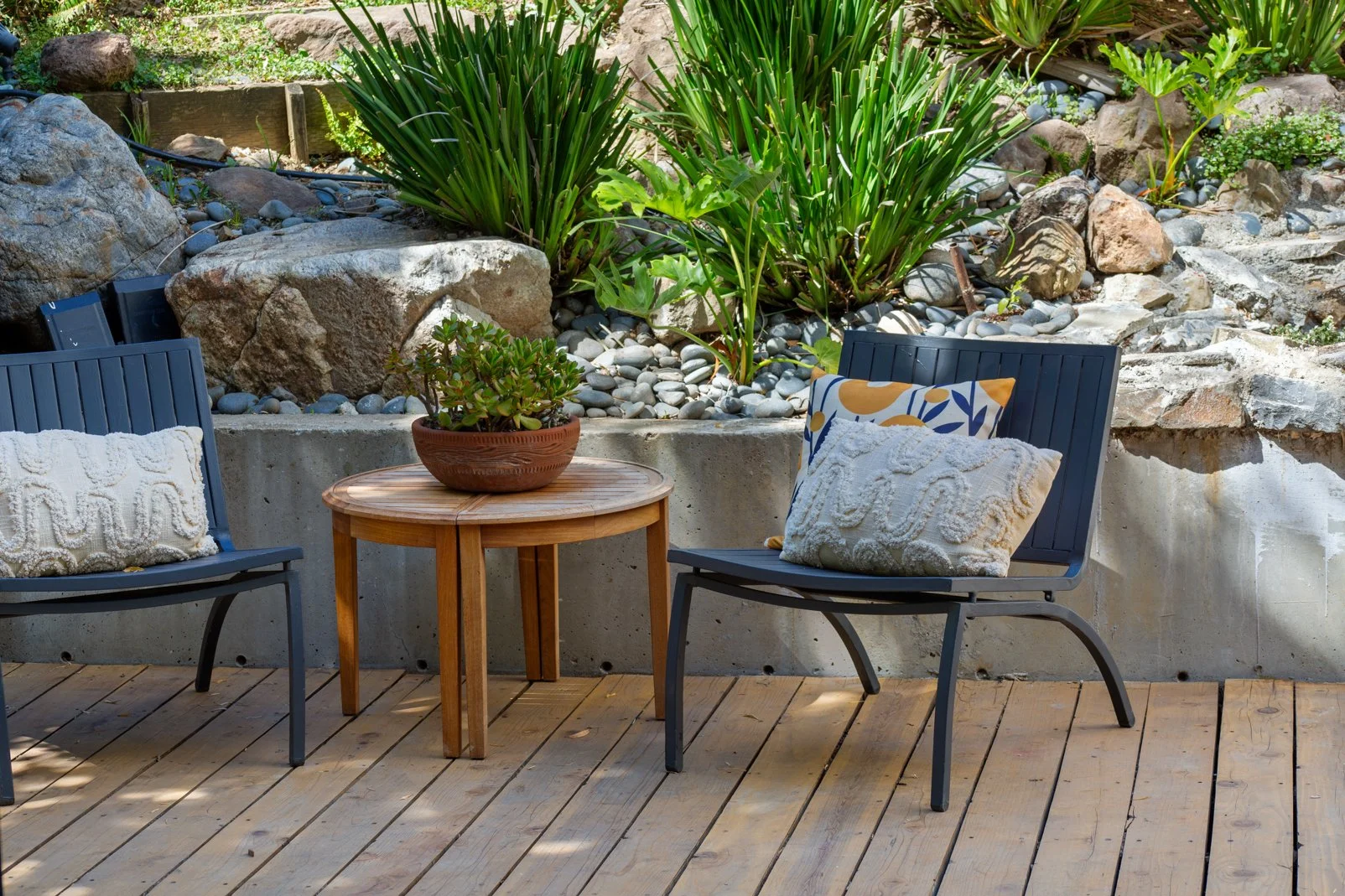 A backyard patio with two black metal chairs decorated with pillows, a small round wooden table with a potted plant, and a rocky garden with green plants and large rocks in the background.