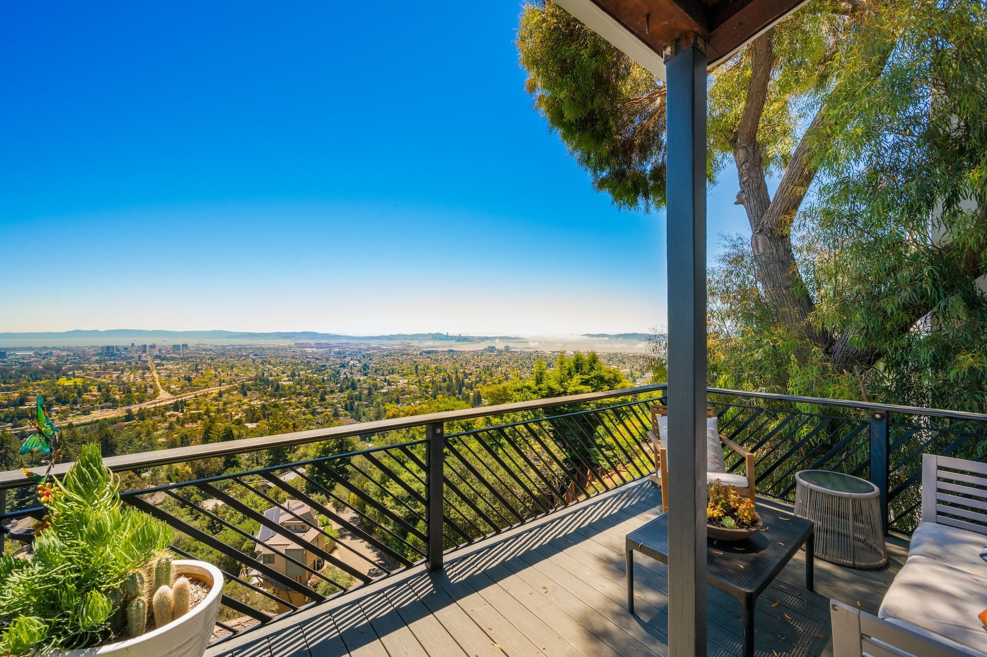 An outdoor balcony with potted succulents, a black metal railing, and a view of a cityscape under a clear blue sky. There are trees and distant buildings visible.