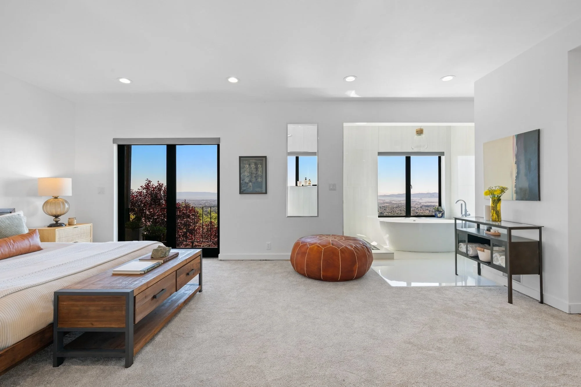 Living room with large windows showing a city view, modern decor, a low wooden coffee table, orange pouf, and a bed on the left with a beige and gray color scheme.
