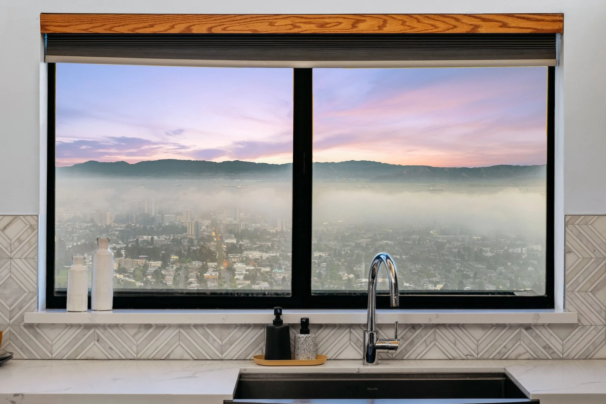 Kitchen countertop with a sink, soap dispenser, and a window showing a cityscape and distant mountains during sunrise or sunset.