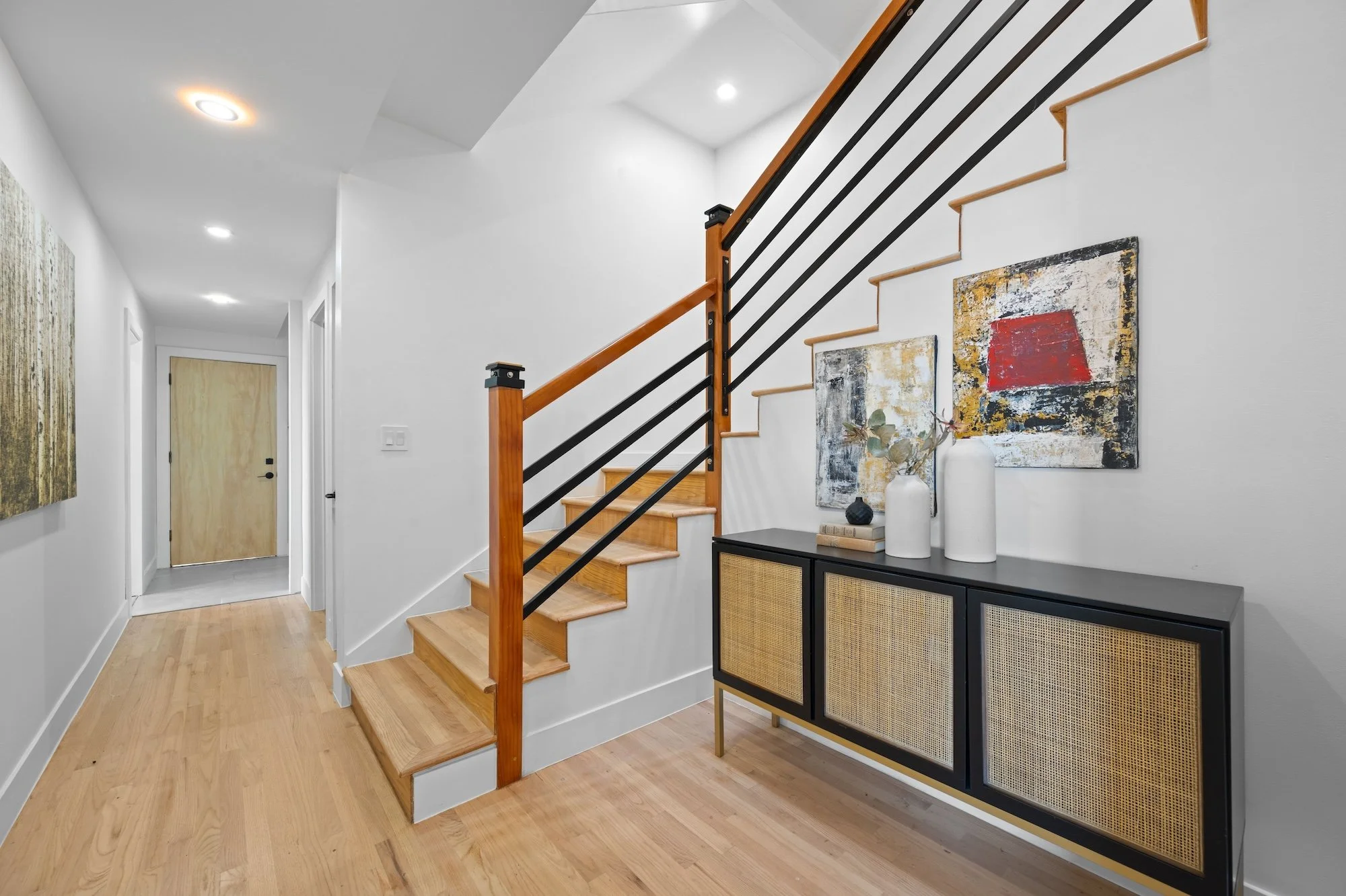 Interior view of a modern home entryway with a staircase, abstract art on the wall, and a black sideboard decorated with vases and a plant.