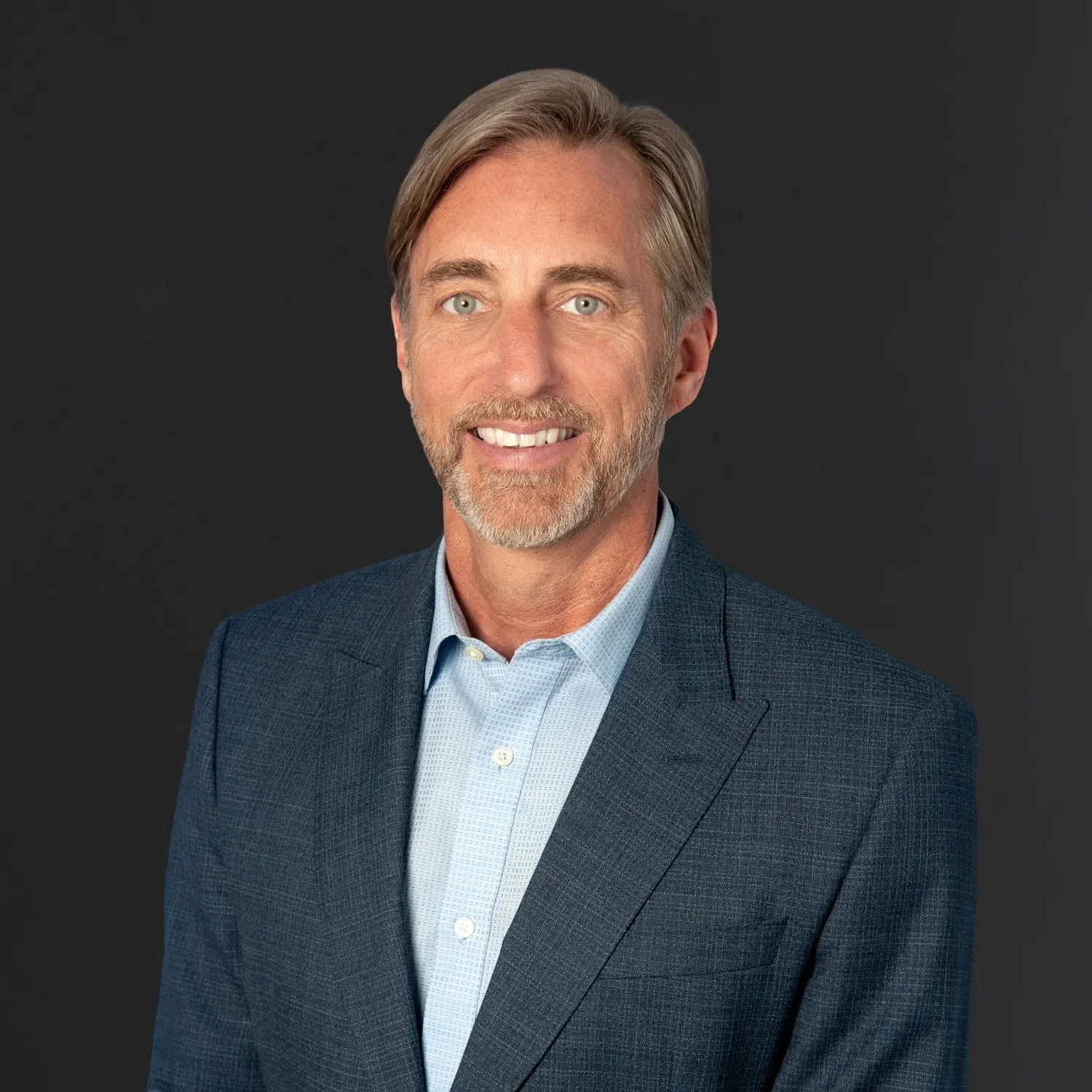 A middle-aged man with light skin, gray hair, blue eyes, and a beard, wearing a dark blue suit jacket and a light blue dress shirt, smiling against a dark gray background.