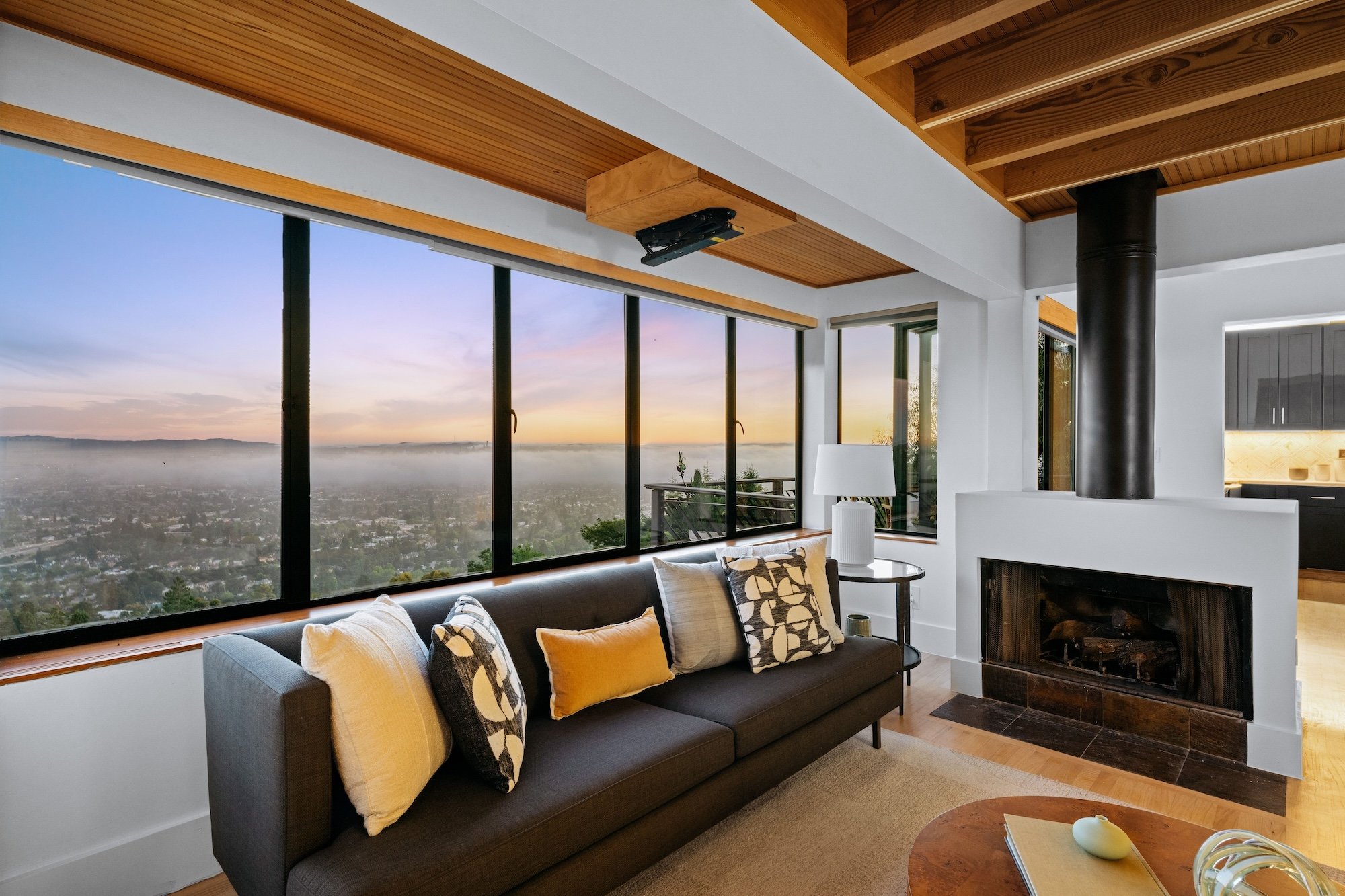 Modern living room with large floor-to-ceiling windows showing a city view at sunset, dark-colored sofa with patterned and yellow pillows, wooden ceiling beams, fireplace, and a coffee table.