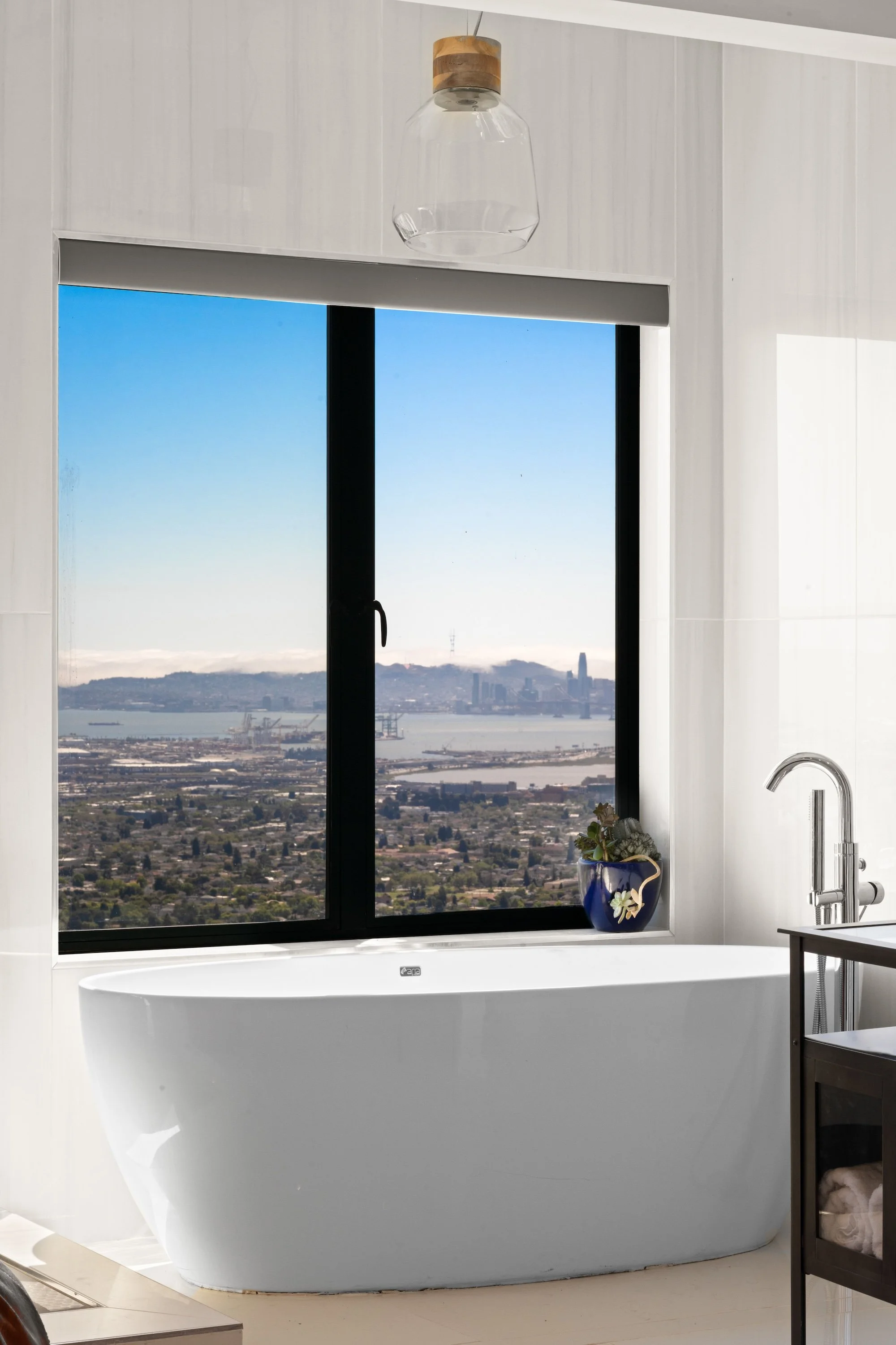 Modern bathroom with a white freestanding bathtub, large window with a view of a city and bay, blue sky, and a village of houses. There is a potted plant on the window sill, a chandelier hanging from the ceiling, and a dark wood side table.