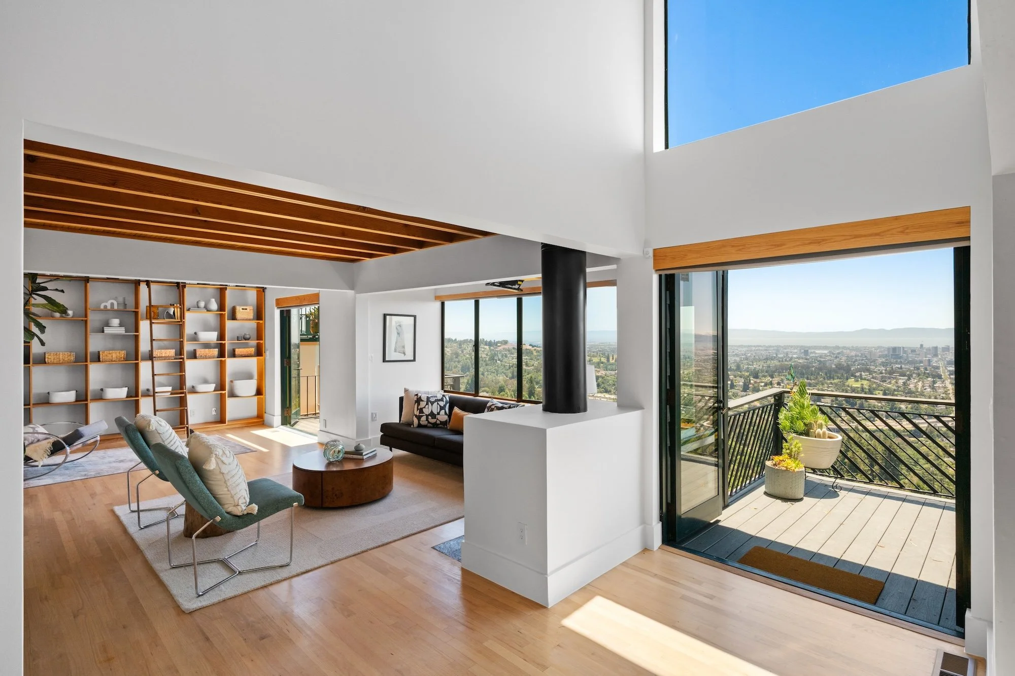 Living room with large windows and sliding glass doors leading to a balcony, modern furniture, built-in wooden shelves, and wood ceiling accents.