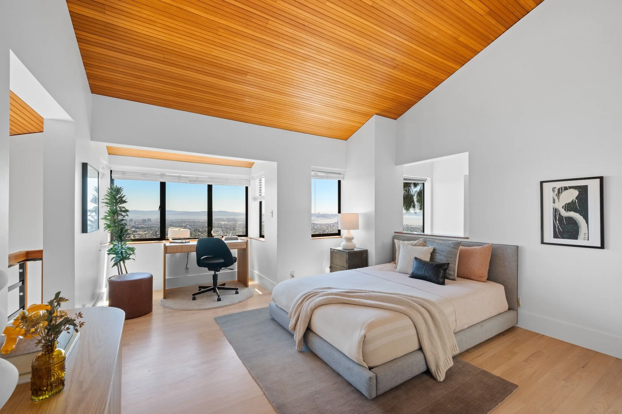 A spacious bedroom with a beige bed, white walls, wooden ceiling, large windows with city and water views, a small desk with a chair, and decorative plants.